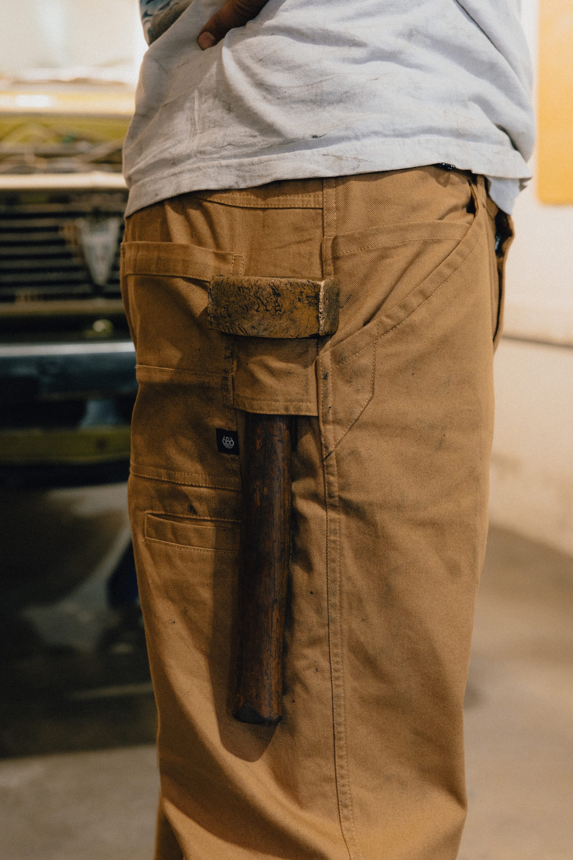 A person wearing 686 Men's Unwork® Tradesman Pant - Relaxed Fit by 686, in tan, with a rusty hammer tucked into the side pocket stands near a blurred car in a garage or workshop.