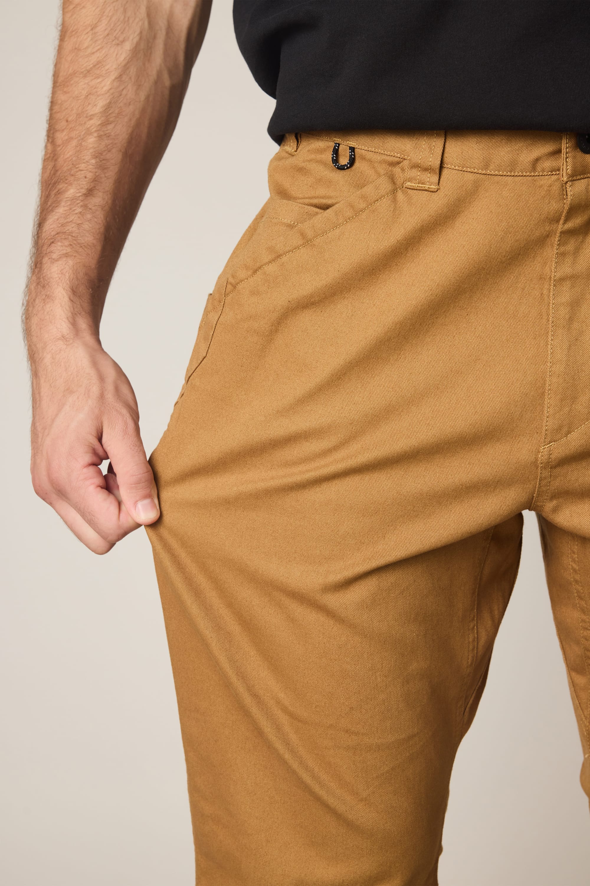 A person wearing the 686 Men's Unwork® Tradesman Pant - Relaxed Fit in tan and a black shirt stretches the nylon woven fabric with one hand, highlighting the pant's flexibility.