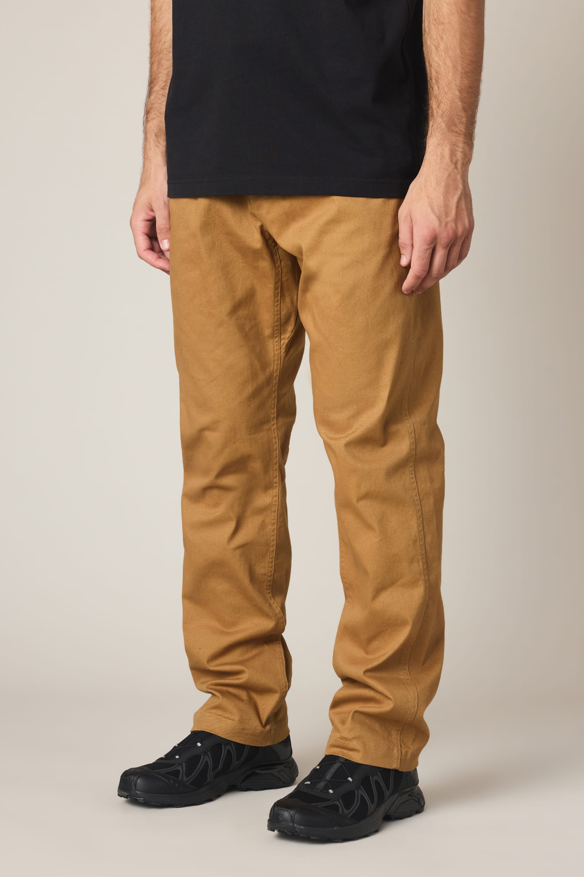 A person wearing 686 Men's Unwork® Tradesman Pant - Relaxed Fit in tan, a black T-shirt, and black sneakers stands against a plain light background. The photo is cropped from the shoulders down.