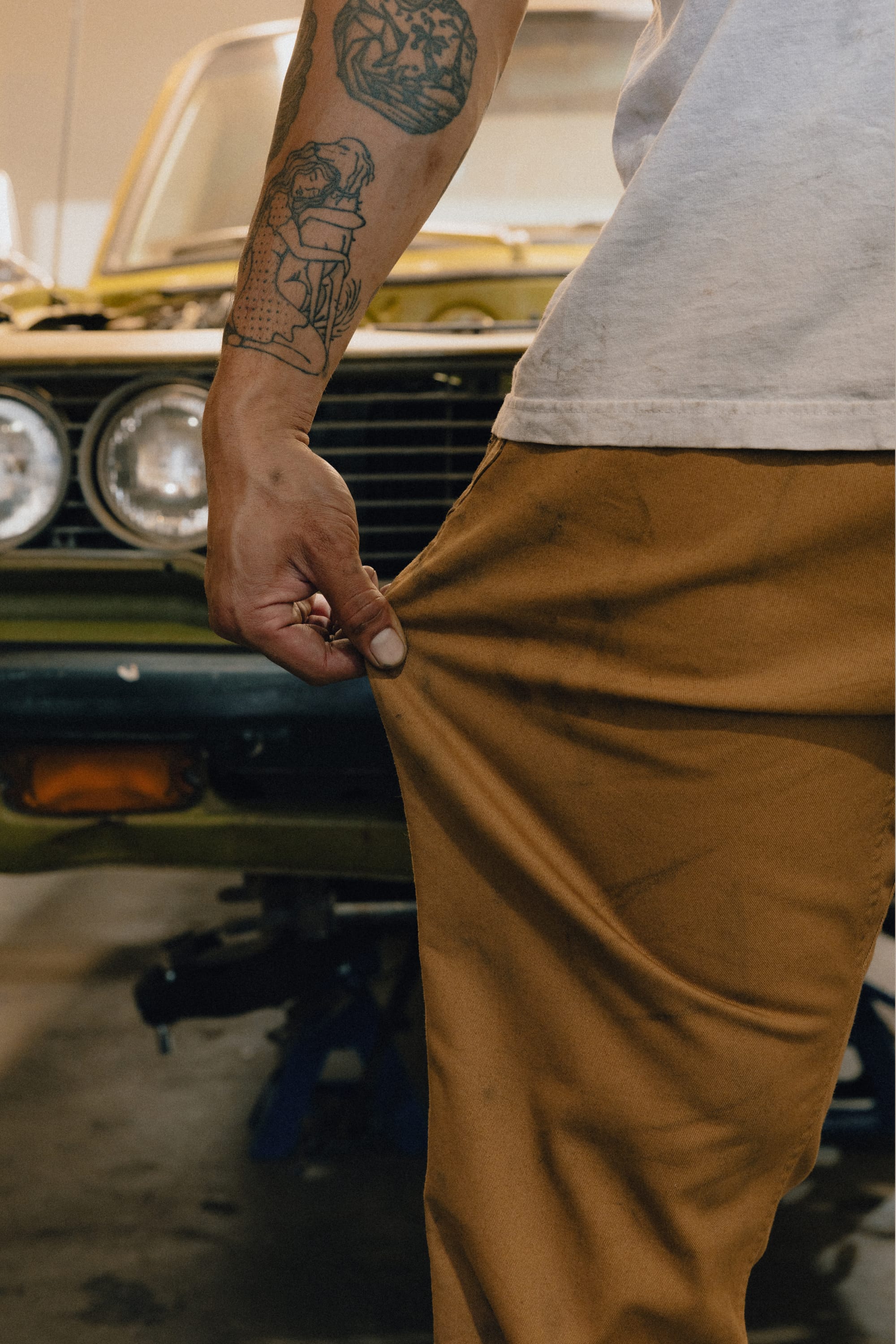 A person with tattoos pinches their 686 Men's Unwork® Tradesman Pant - Relaxed Fit while standing near an olive green vintage car. Only the person's arm, torso, and hip are visible in the image.