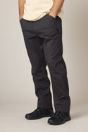 A person stands with hands in pockets wearing 686 Men's Unwork® Tradesman Pant - Relaxed Fit in dark gray against a plain background. The image is cropped from the shoulders down.