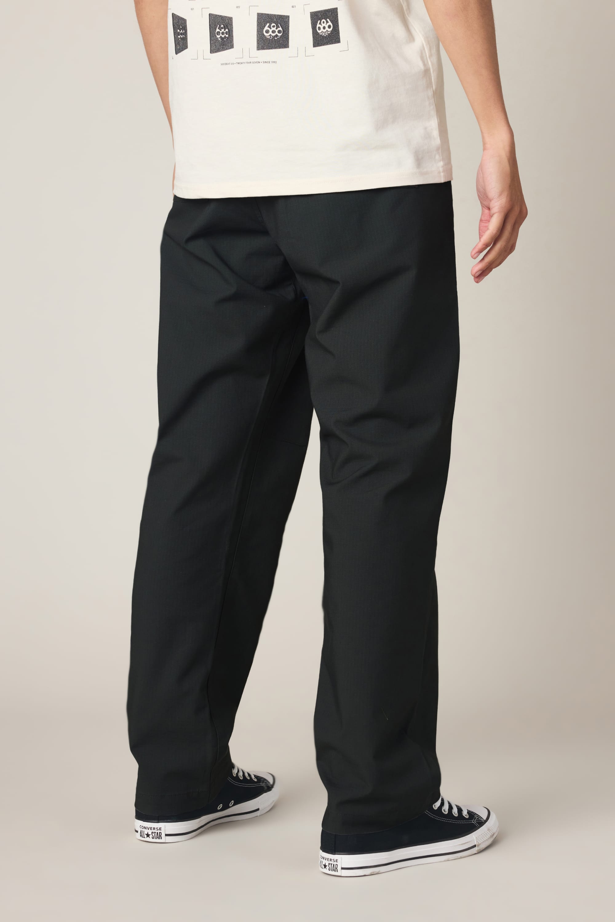 A person stands facing away, wearing a white graphic T-shirt, the 686 Men's Free Loader Pant - Loose Fit by 686 in black spandex woven fabric, and black low-top sneakers with white soles on a plain light background.