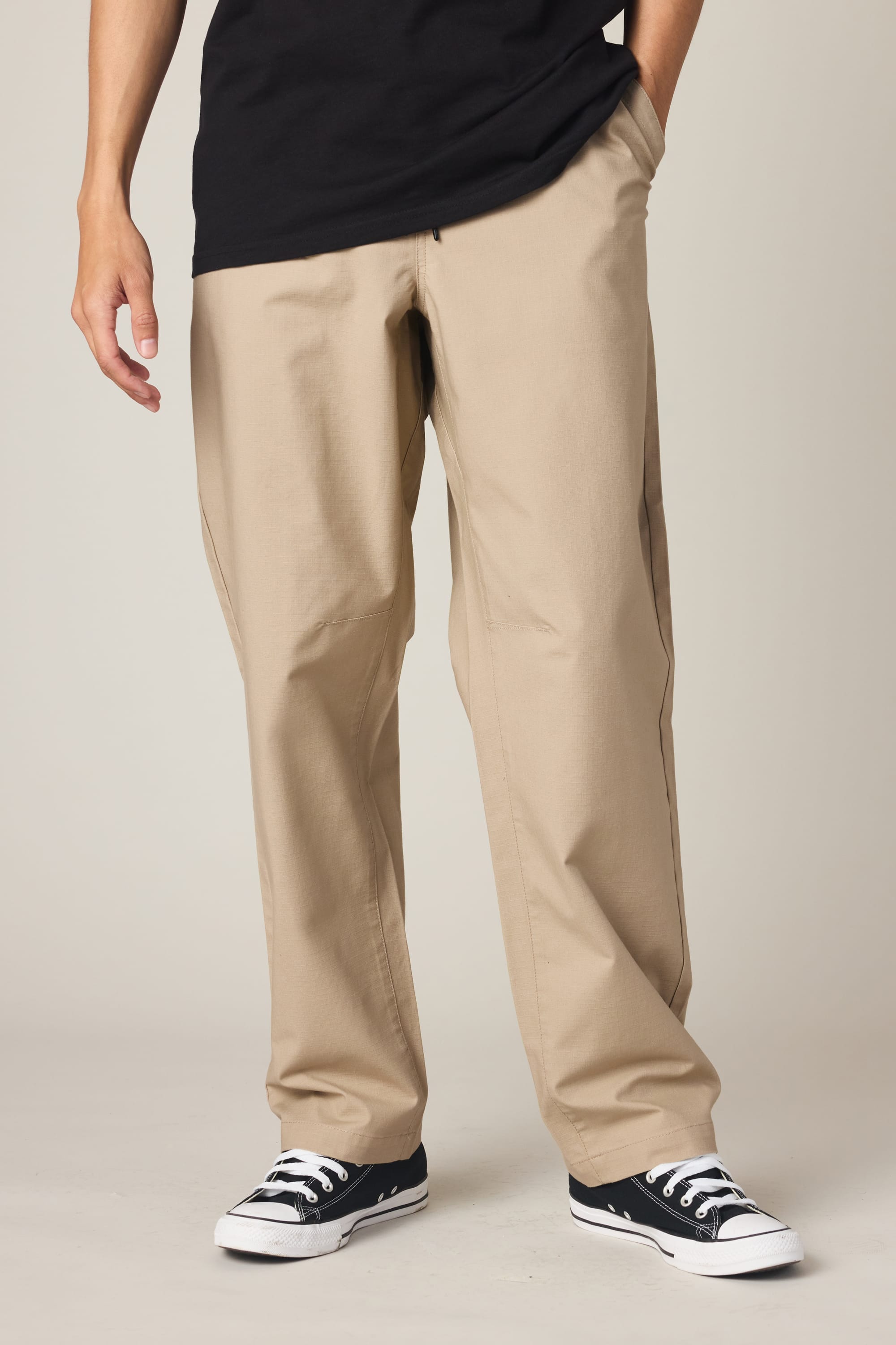 A person wearing a black T-shirt, the 686 Men’s Free Loader Pant - Loose Fit by 686 in beige, and black-and-white sneakers stands with one hand in their pocket against a plain background.