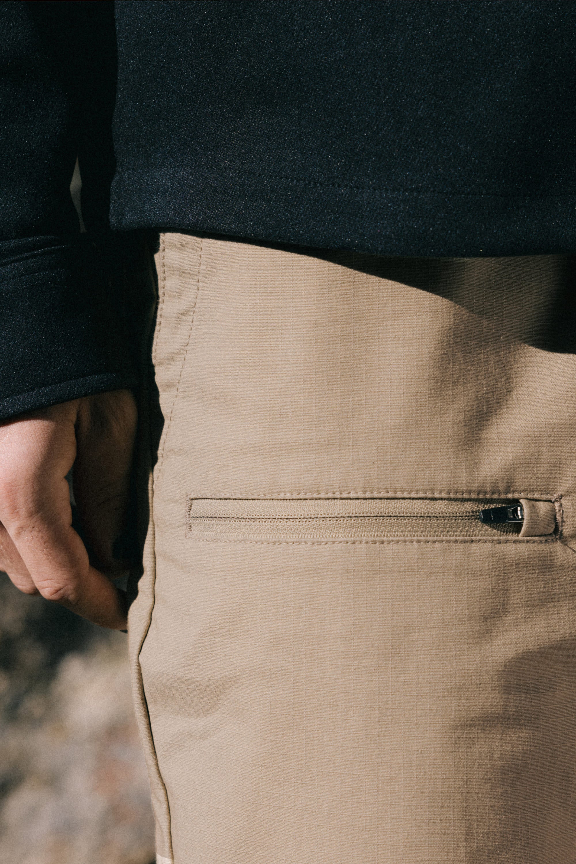 A close-up of a person wearing the 686 Men's Free Loader Pant - Loose Fit in tan with a zippered pocket, paired with a black long-sleeve top, showing part of their left hand and side.