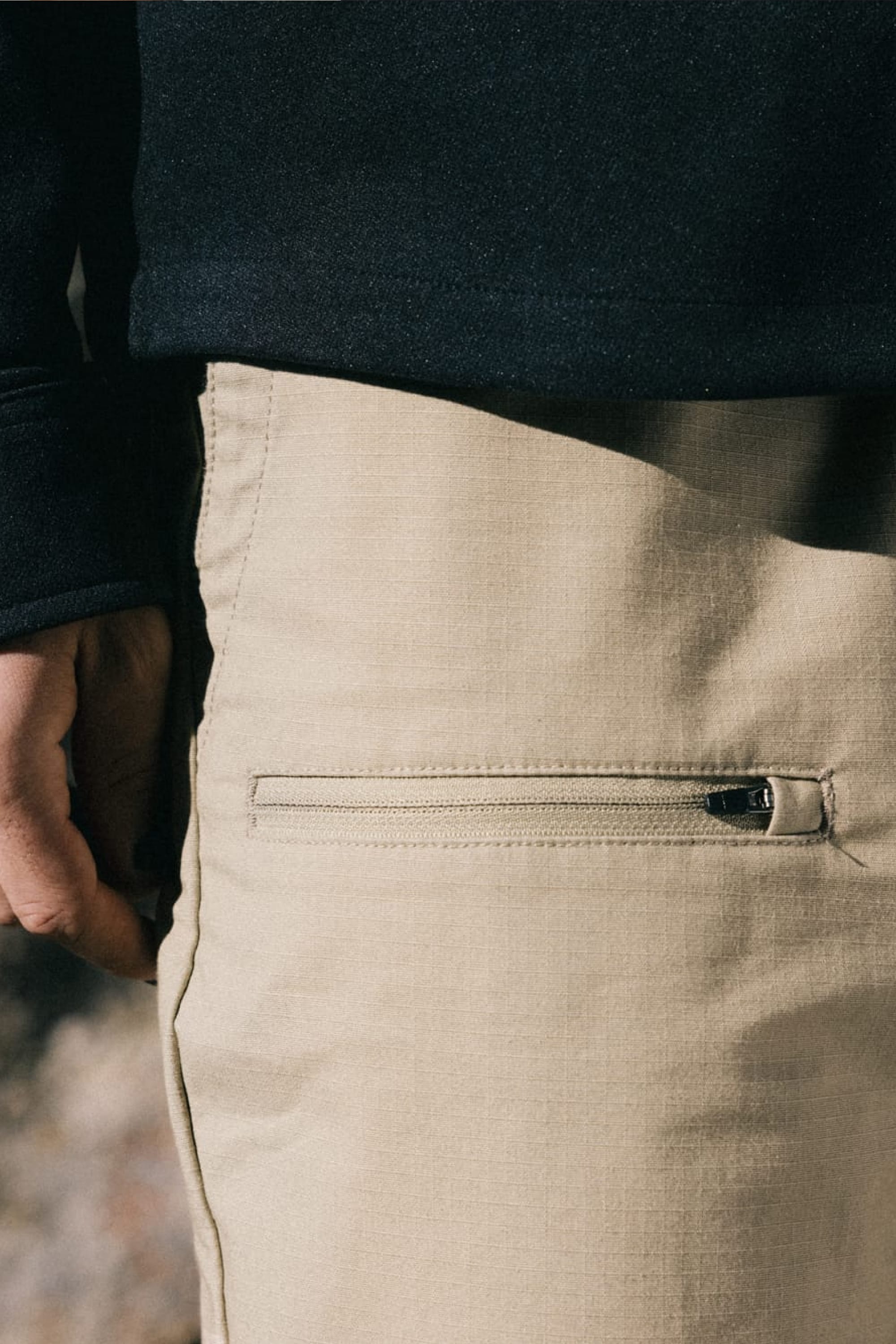 A close-up of a person’s hand by their side, wearing the 686 Men’s Free Loader Pant - Loose Fit in light beige with a zippered pocket, paired with a black long-sleeve shirt.