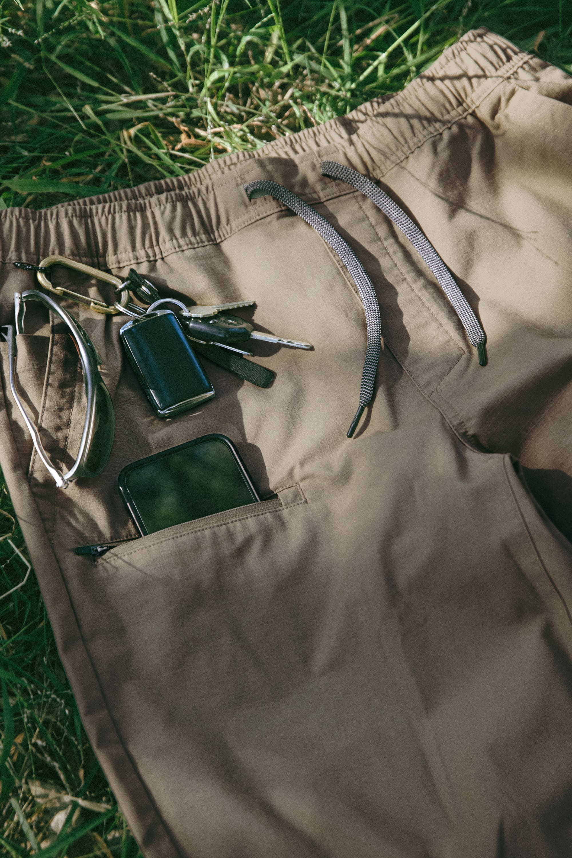 The 686 Men’s Free Loader Pant - Loose Fit in beige is displayed lying on grass, with a keychain, car key, and smartphone placed on top and partially tucked into a zippered pocket.