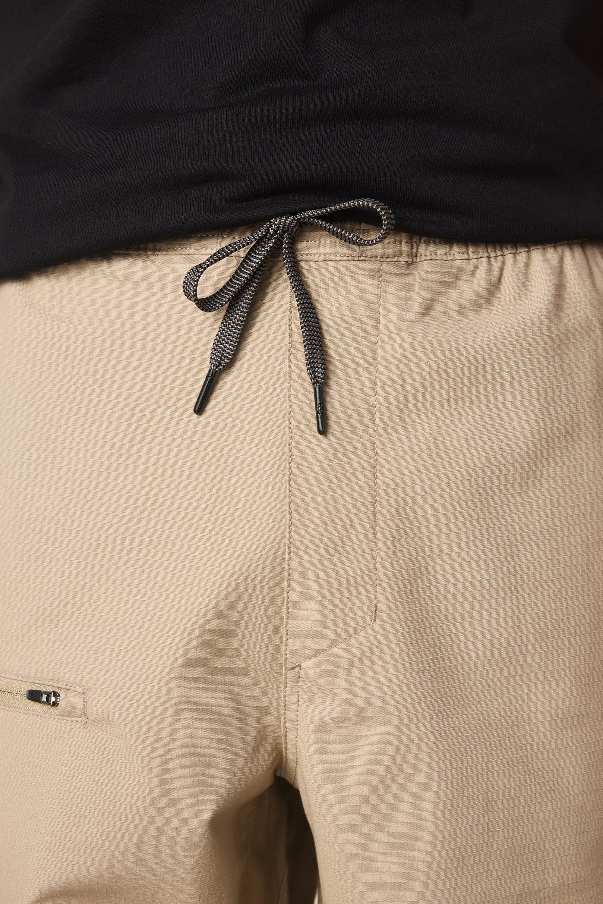 A close-up of a person wearing the 686 Men’s Free Loader Pant - Loose Fit in beige, featuring a visible drawstring tied in a bow and paired with a black shirt. The nylon woven design by 686 includes a small zippered pocket on the left side for added functionality.