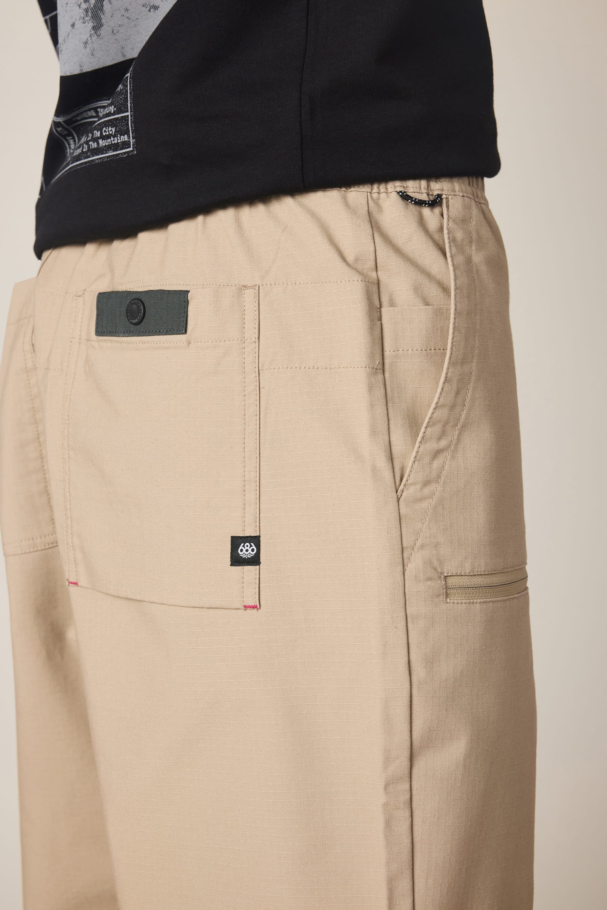 Close-up of a person wearing the 686 Men’s Free Loader Pant - Loose Fit by 686, featuring tan cargo styling inspired by premium men’s pants, detailed with a black buttoned pocket and a small black tag, paired with a black shirt. The focus is on the pants’ meticulous stitching and standout design elements.