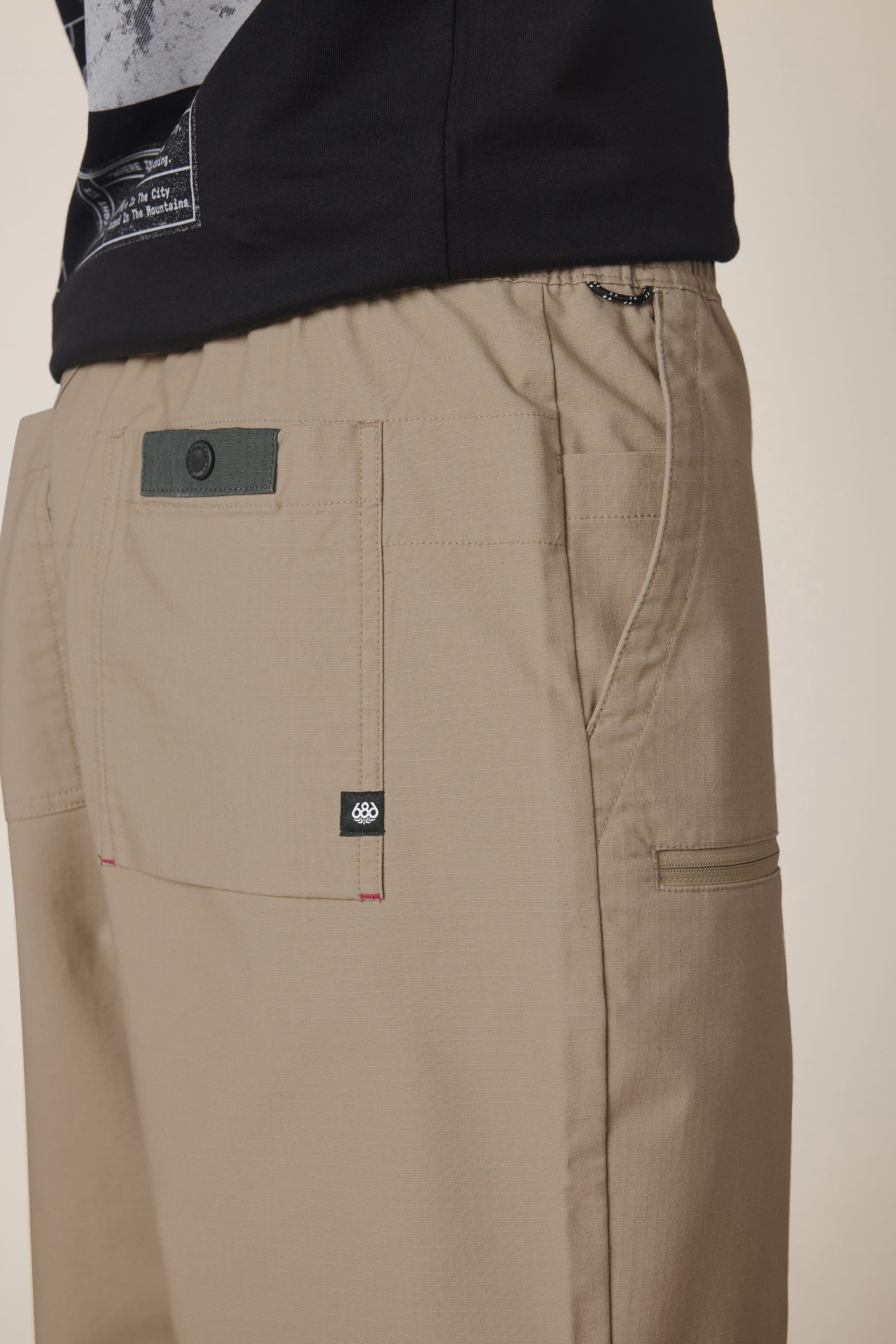 Close-up of a person wearing the 686 Men's Free Loader Pant - Loose Fit in beige, featuring visible front and back pockets, a small black tag, and a black shirt tucked in at the waist.