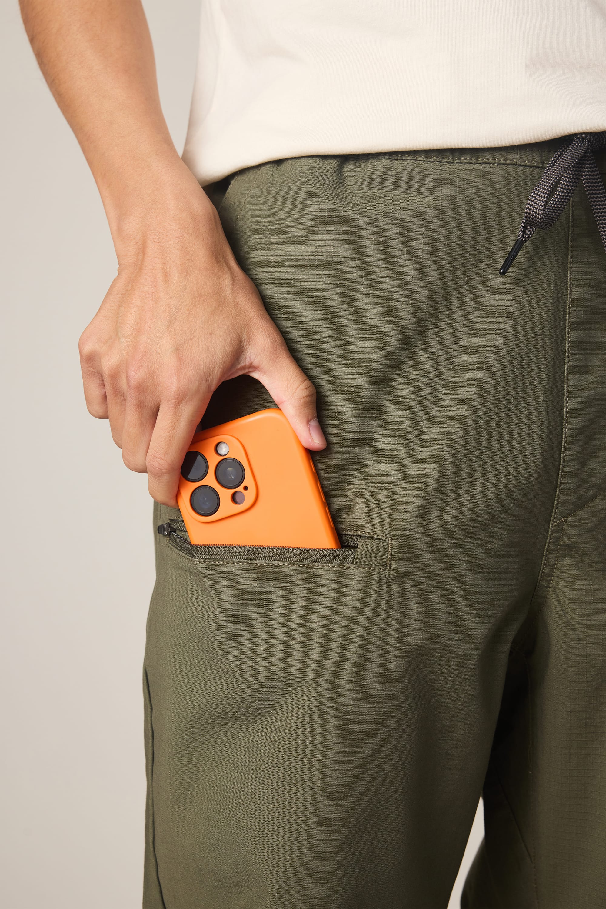 A man in 686 Men's Free Loader Pant - Loose Fit in olive green and a white shirt slips an orange smartphone with black camera lenses into the side pocket.