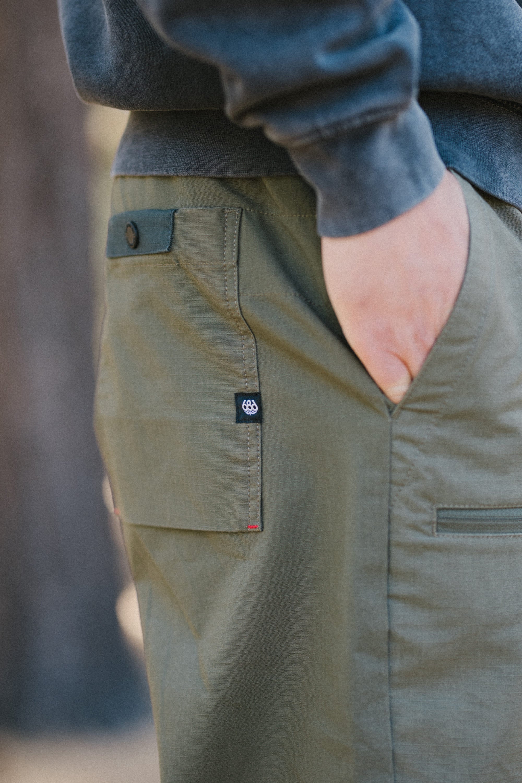 Close-up of a person wearing the 686 Men's Free Loader Pant - Loose Fit, an olive green nylon woven cargo pant from 686 featuring a flap pocket with a dark button. The person has one hand partially tucked into the pocket and is also wearing a dark long-sleeve shirt.