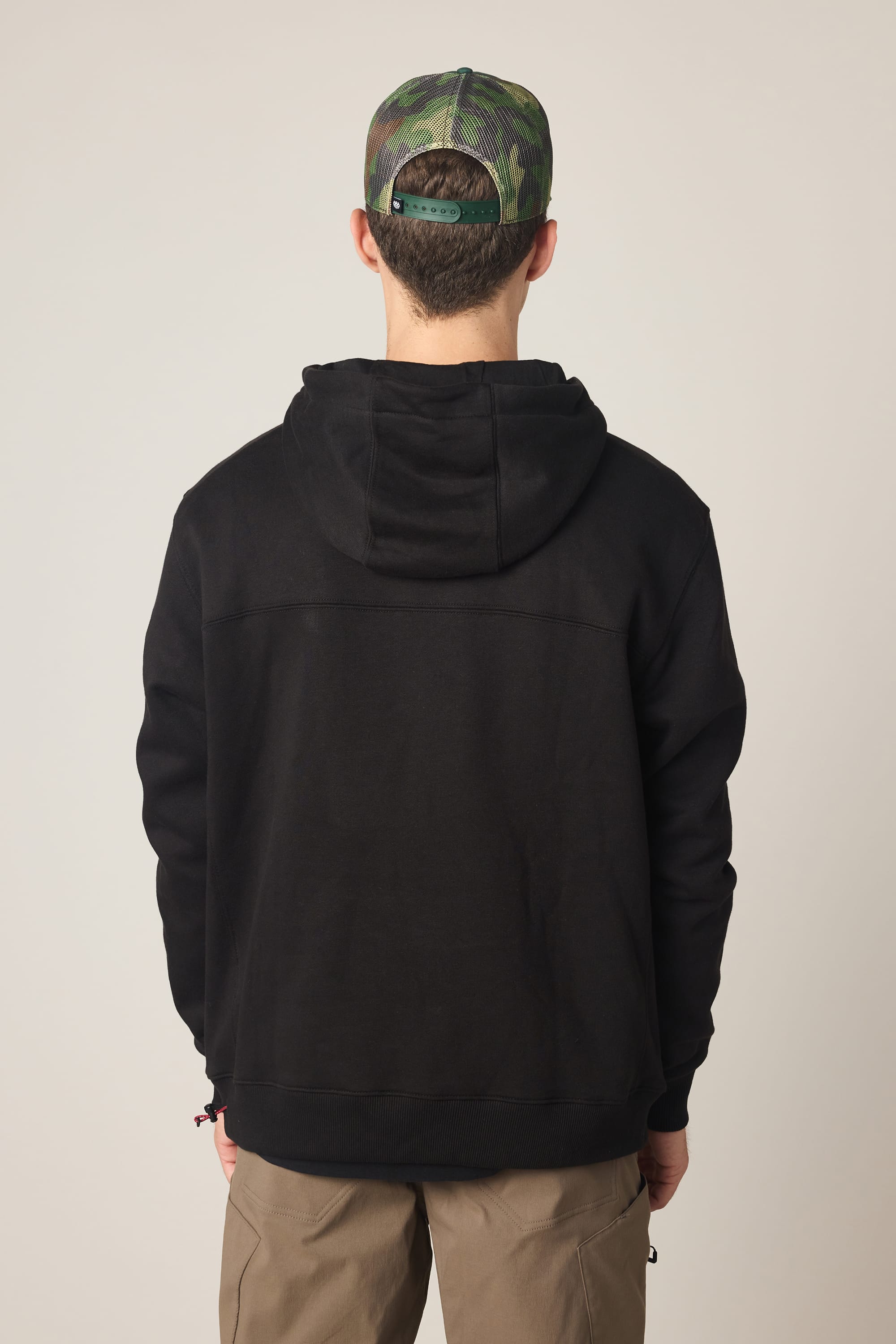 A person stands with their back to the camera, wearing a 686 Men's All Season Hoody in black, beige pants, and a green camouflage baseball cap against a plain light background.