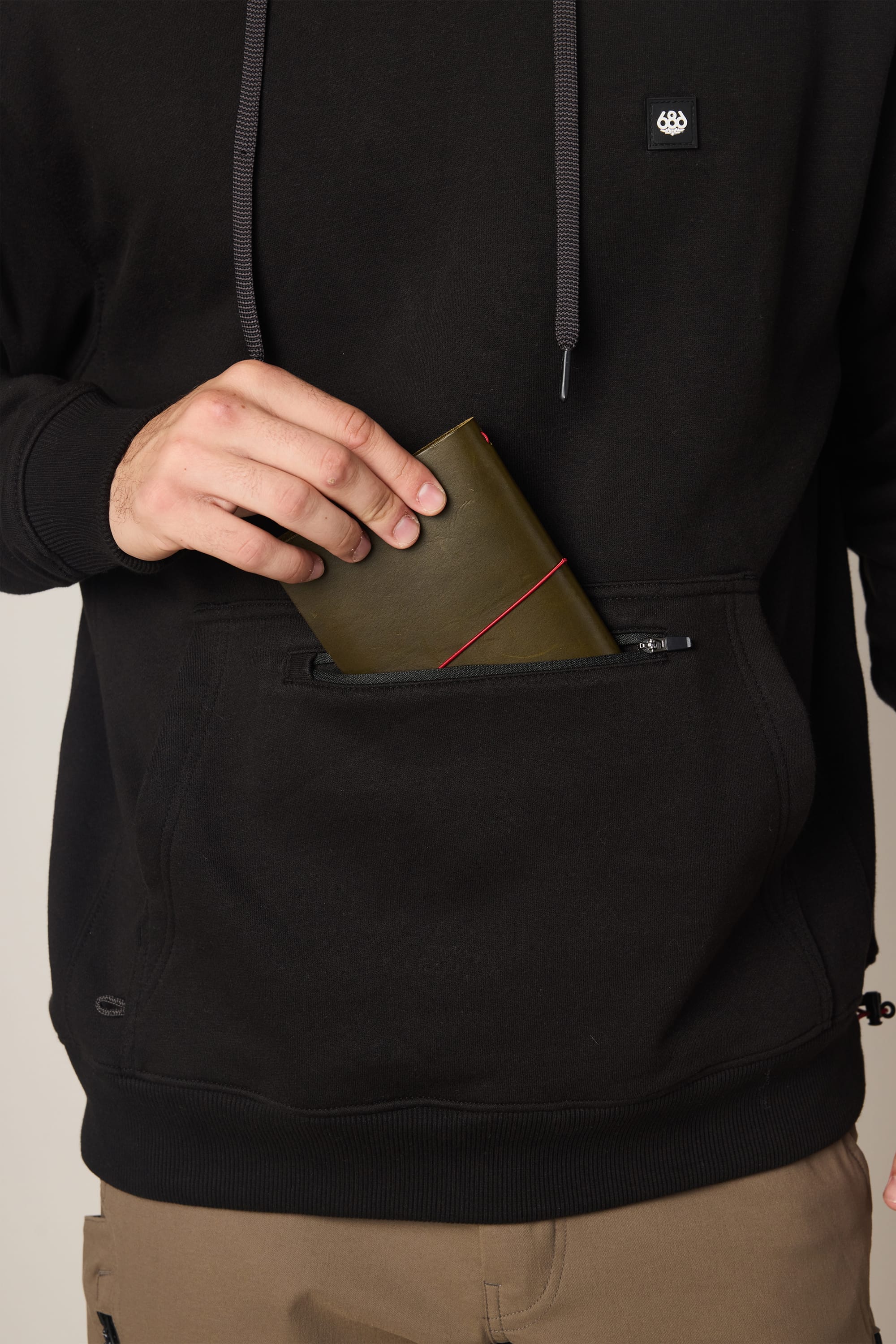 A person wearing a 686 Men's All Season Hoody in black is placing a green wallet with a red elastic band into the front pocket of the hoody. Only their torso and one hand are visible.