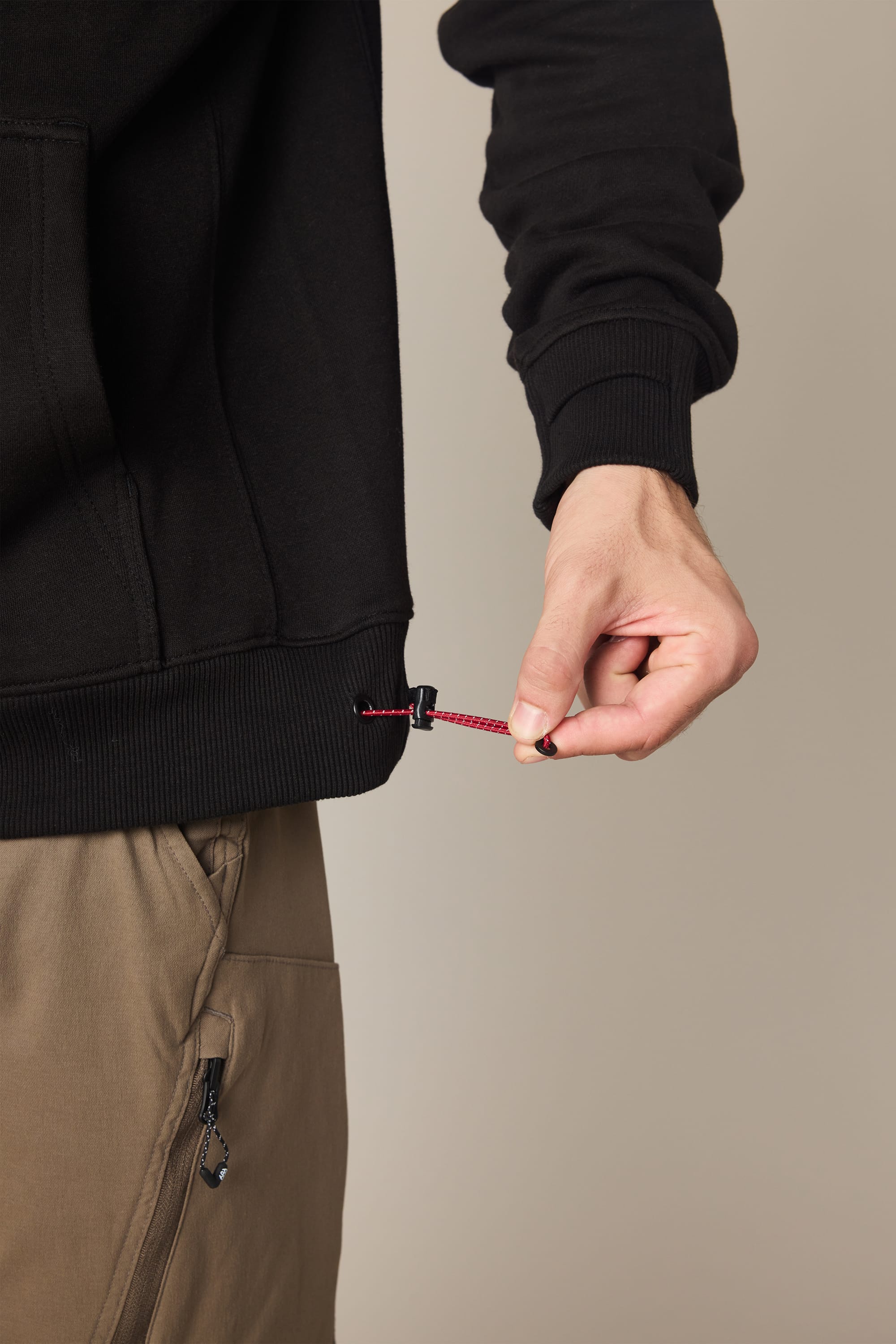 Someone adjusts the red drawstring at the hem of a 686 Men's All Season Hoody in black, paired with tan pants featuring a visible zippered pocket. The plain, neutral background keeps the focus on the outfit.