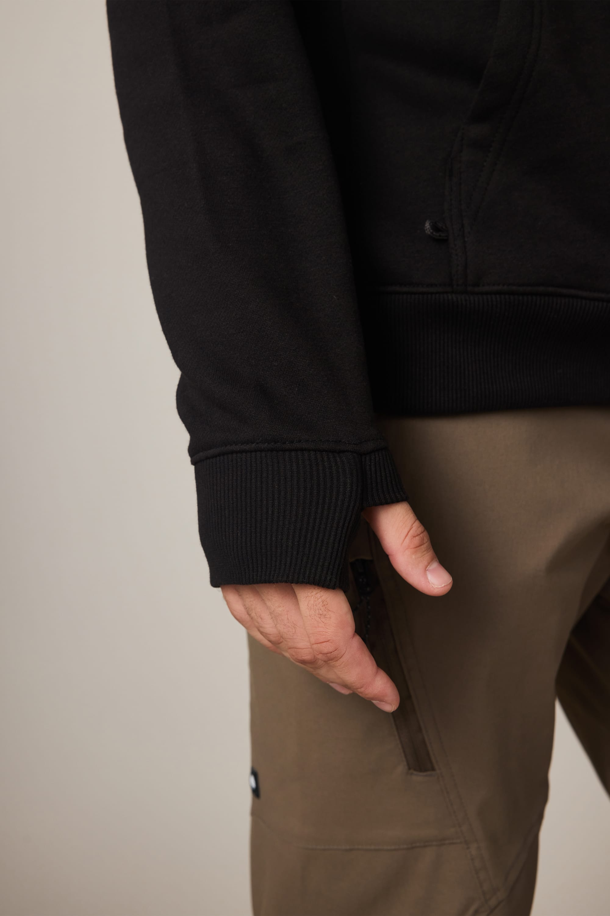 A close-up highlights the sleeve and part of the pants as a person wears the 686 Men's All Season Hoody in black with ribbed cuffs and brown pants, their hand relaxed at their side.