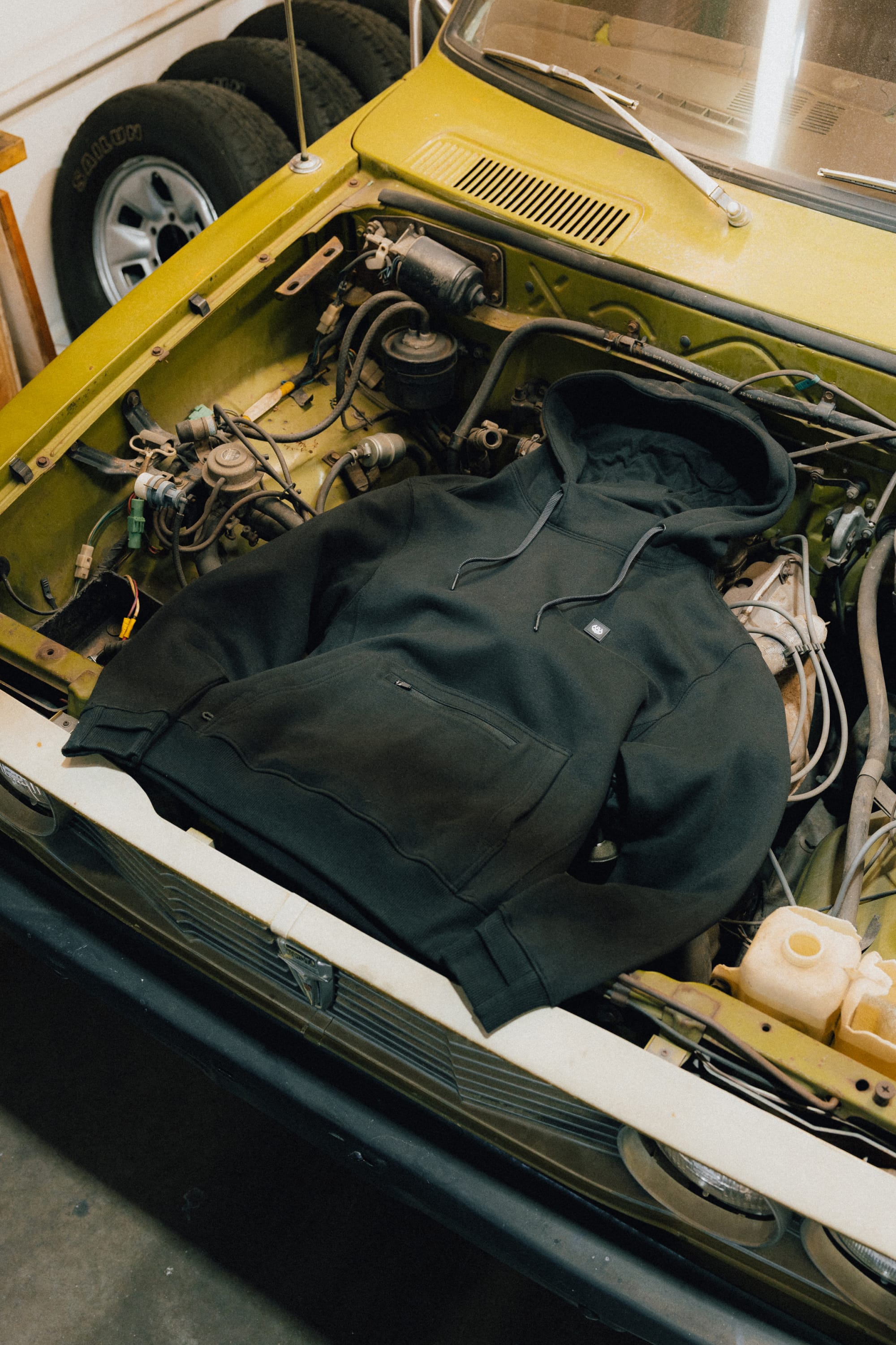 A 686 Men's All Season Hoody in black is laid out over the engine bay of a yellow car with its hood open inside a garage.