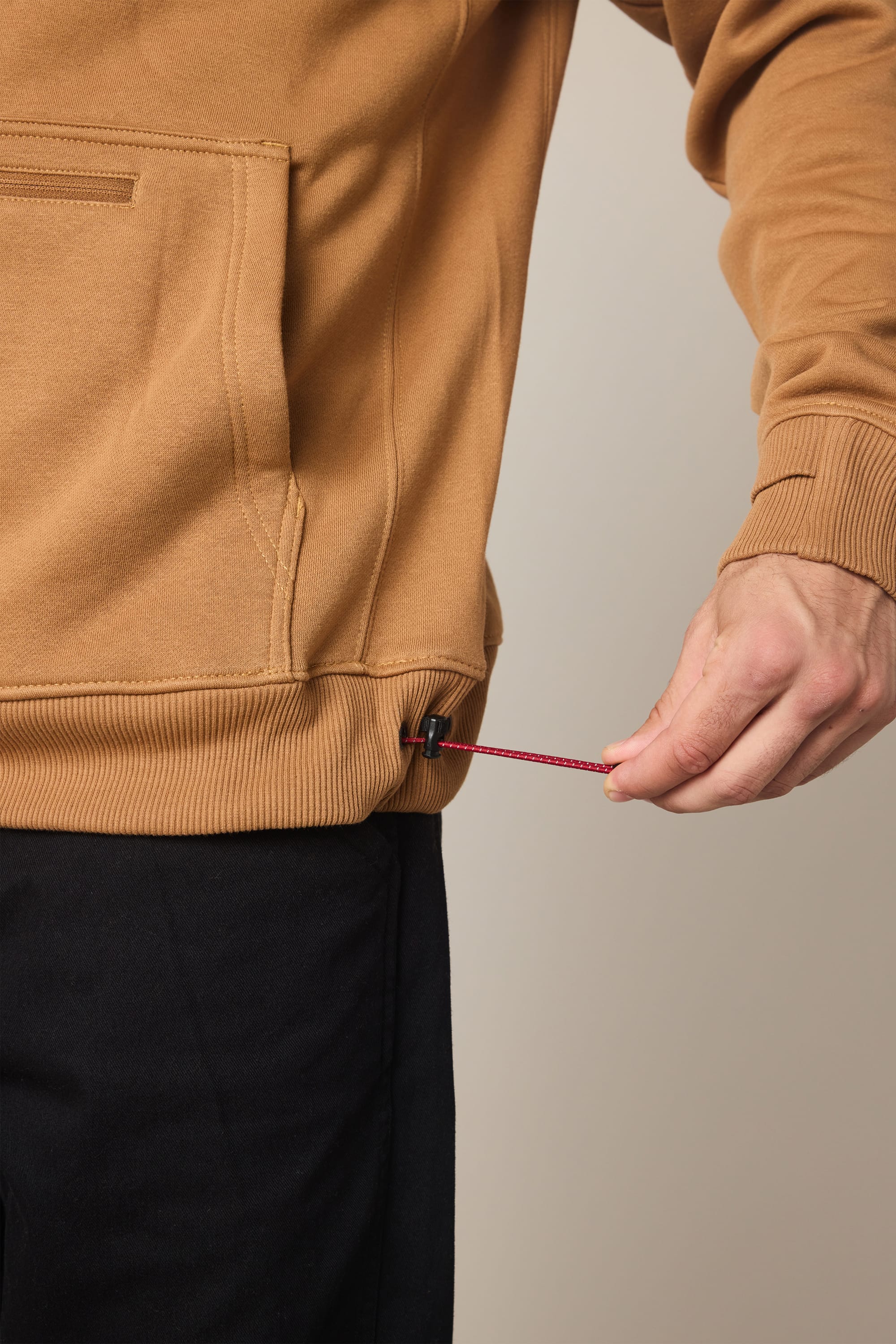 A person wearing the 686 Men's All Season Hoody in brown and black pants adjusts the red drawstring at the bottom hem of the hoodie with their right hand.