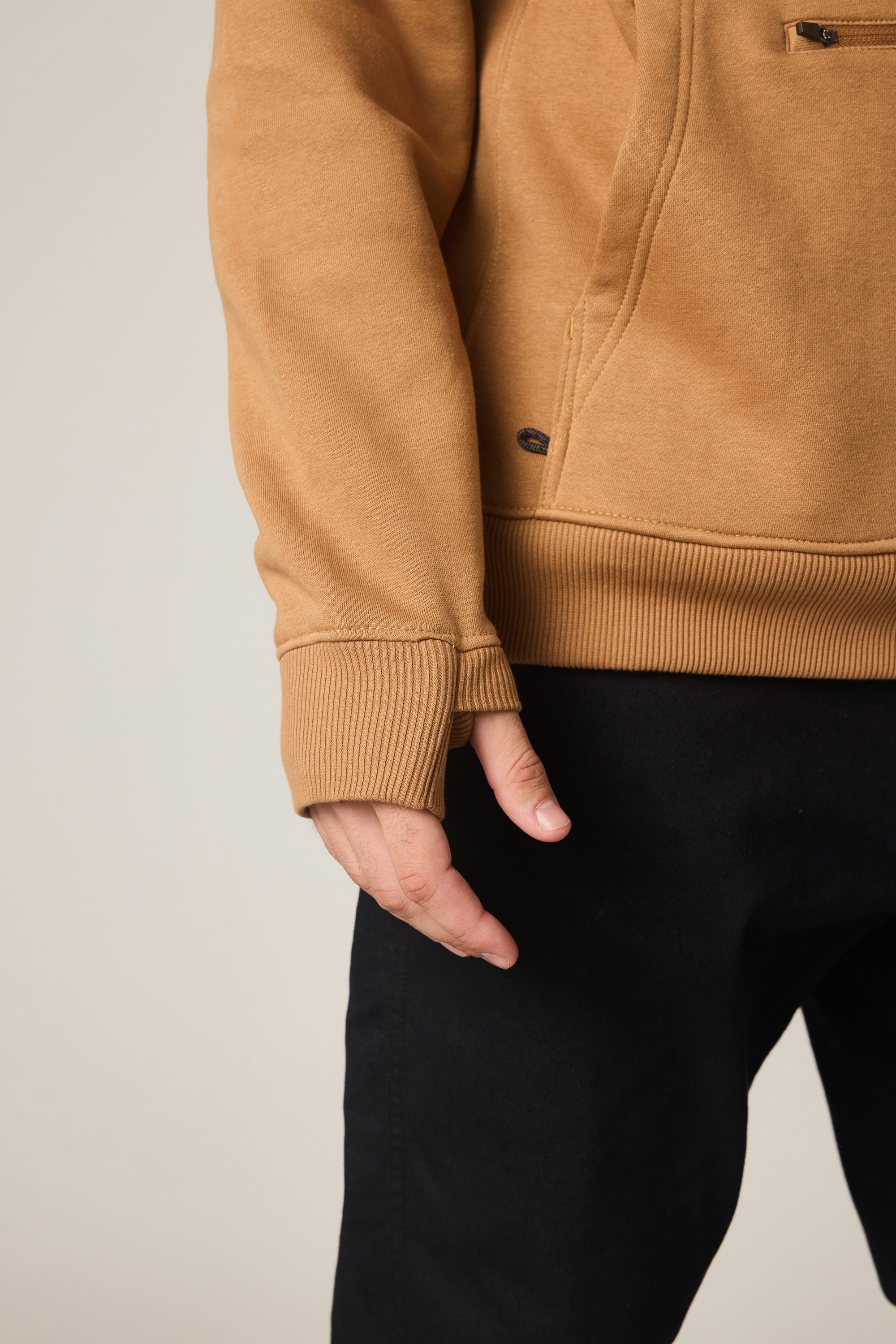 A person wearing the 686 Men's All Season Hoody in tan and black pants stands with their left hand relaxed by their side. The image highlights the lower sleeve of the 686 hoody and hand against a neutral background.