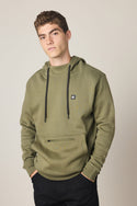 A young man with short curly hair wears the 686 Men's All Season Hoody in olive green with black drawstrings and a small chest logo, paired with black pants, standing hands-in-pocket against a plain light background.