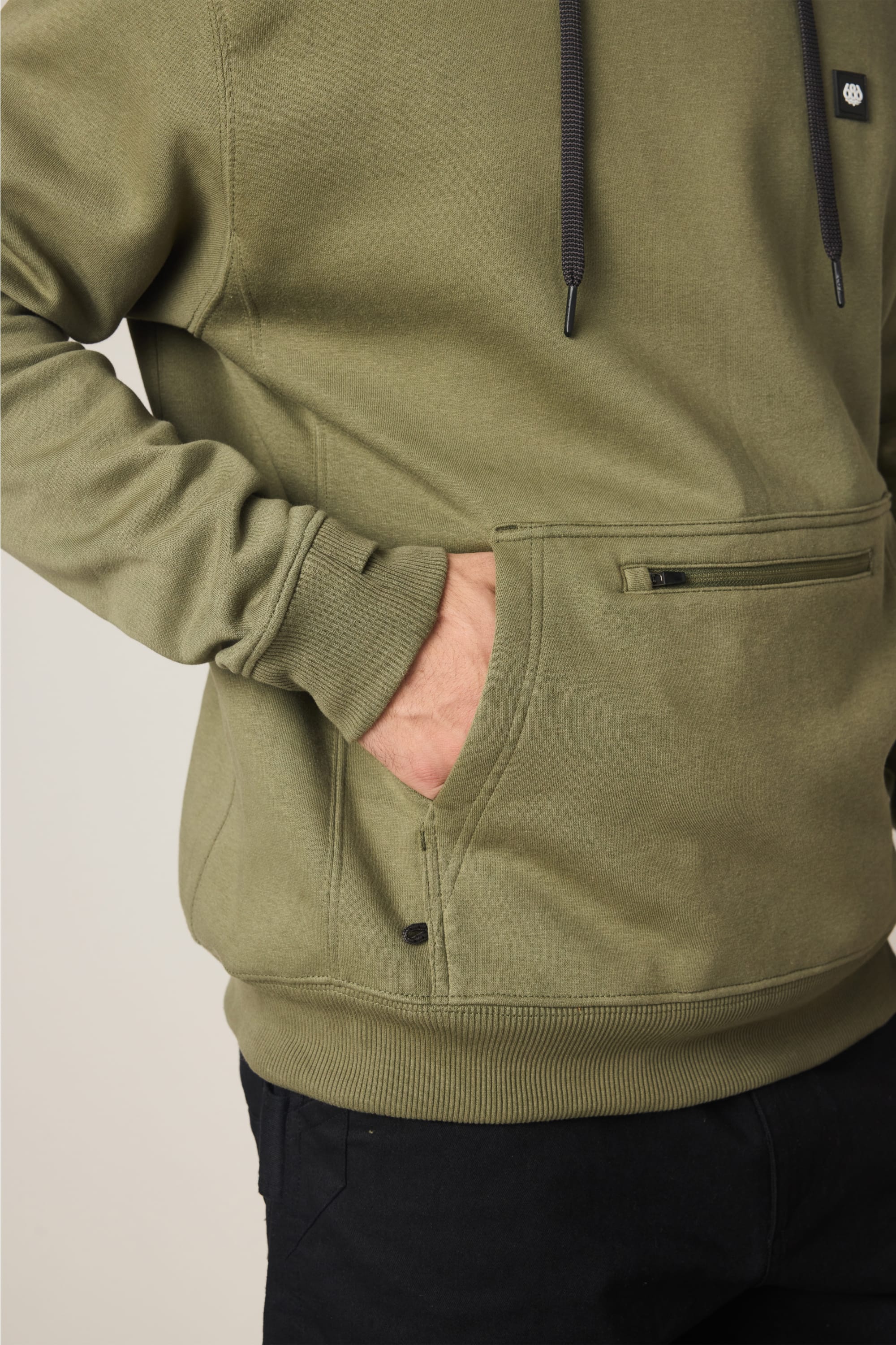 A person wears the 686 Men's All Season Hoody in olive green, featuring a front pocket and black drawstrings, with their right hand in the pocket. The outfit is completed with black pants.