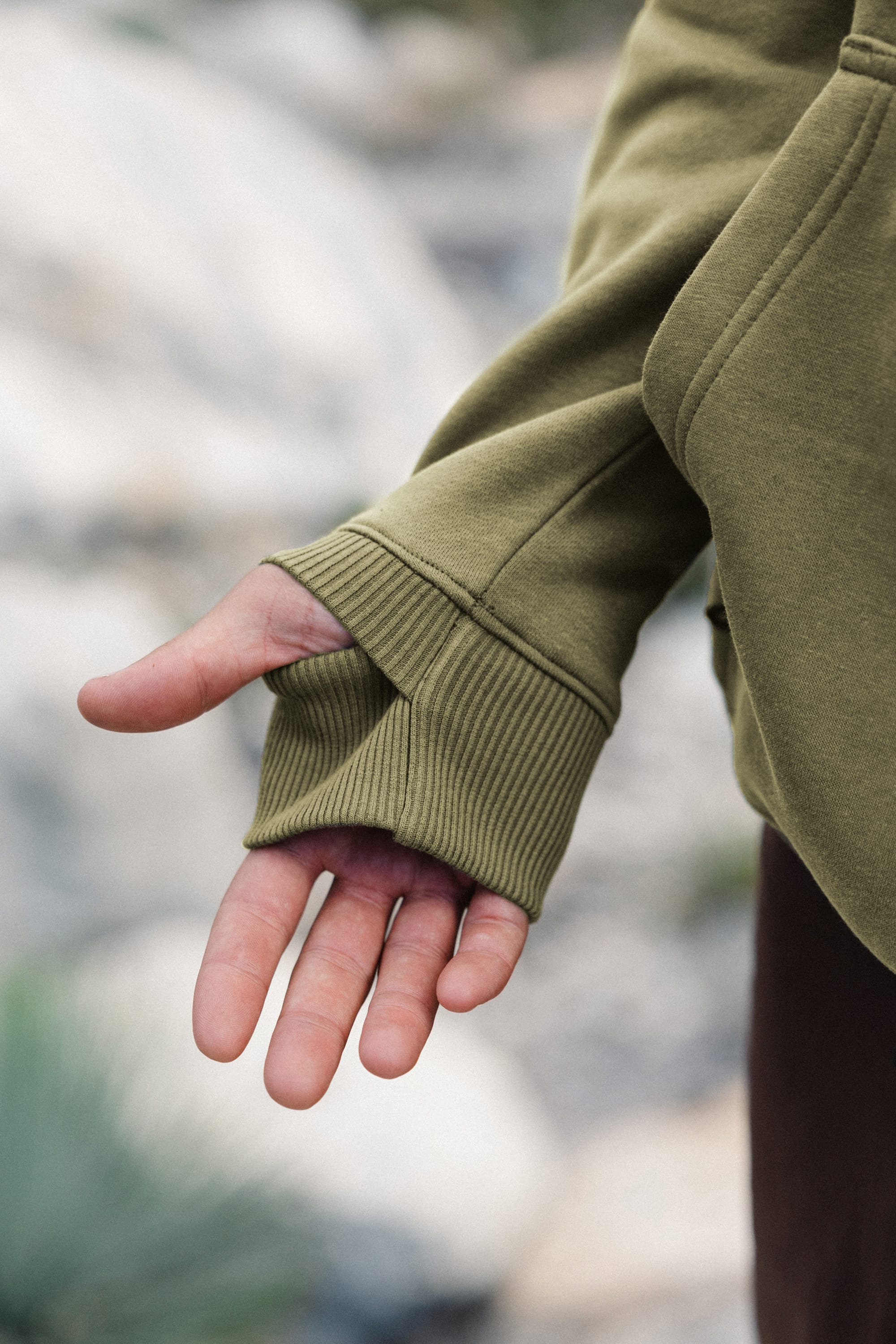 A person’s left hand, partially covered by the sleeve of a 686 Men's All Season Hoody in olive green, with the thumb through a thumb hole. The background is blurred, suggesting an outdoor setting.