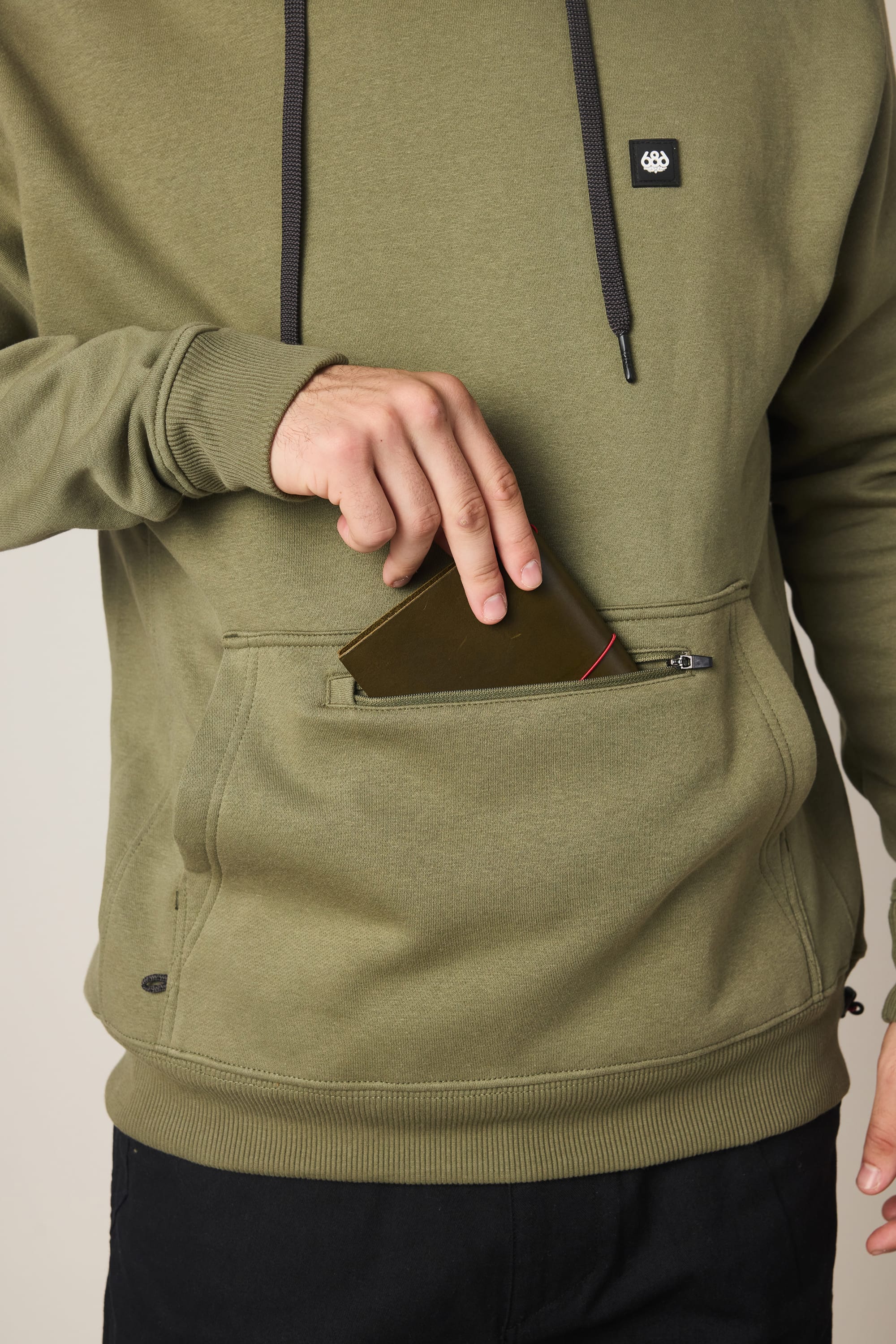 A person wearing the 686 Men's All Season Hoody in olive green places a wallet into its zippered front pocket; only the torso and one hand are visible, with the face not shown.