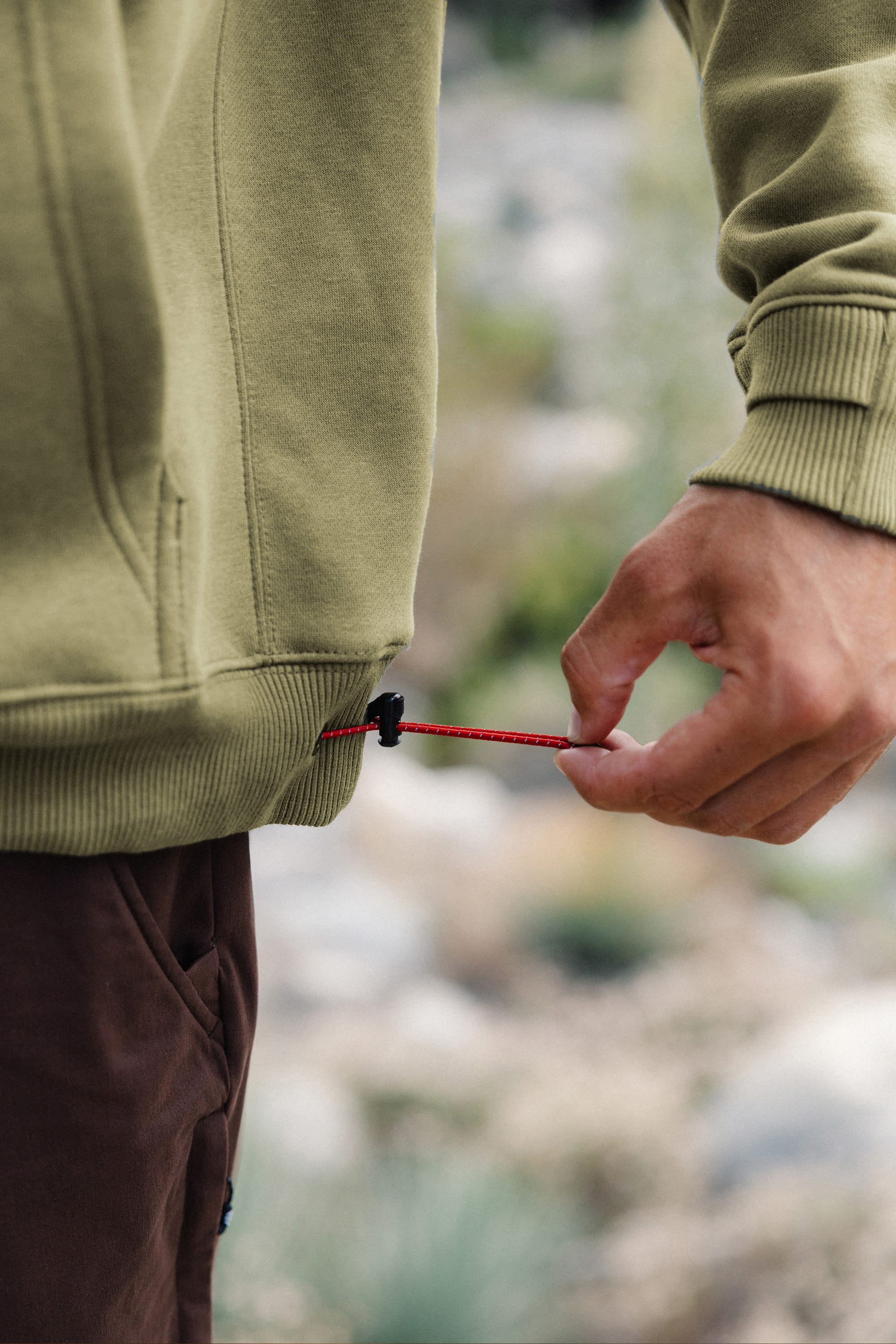 A person adjusts the red drawstring on the hem of a 686 Men's All Season Hoody in olive green while standing outdoors, with a blurred natural background.
