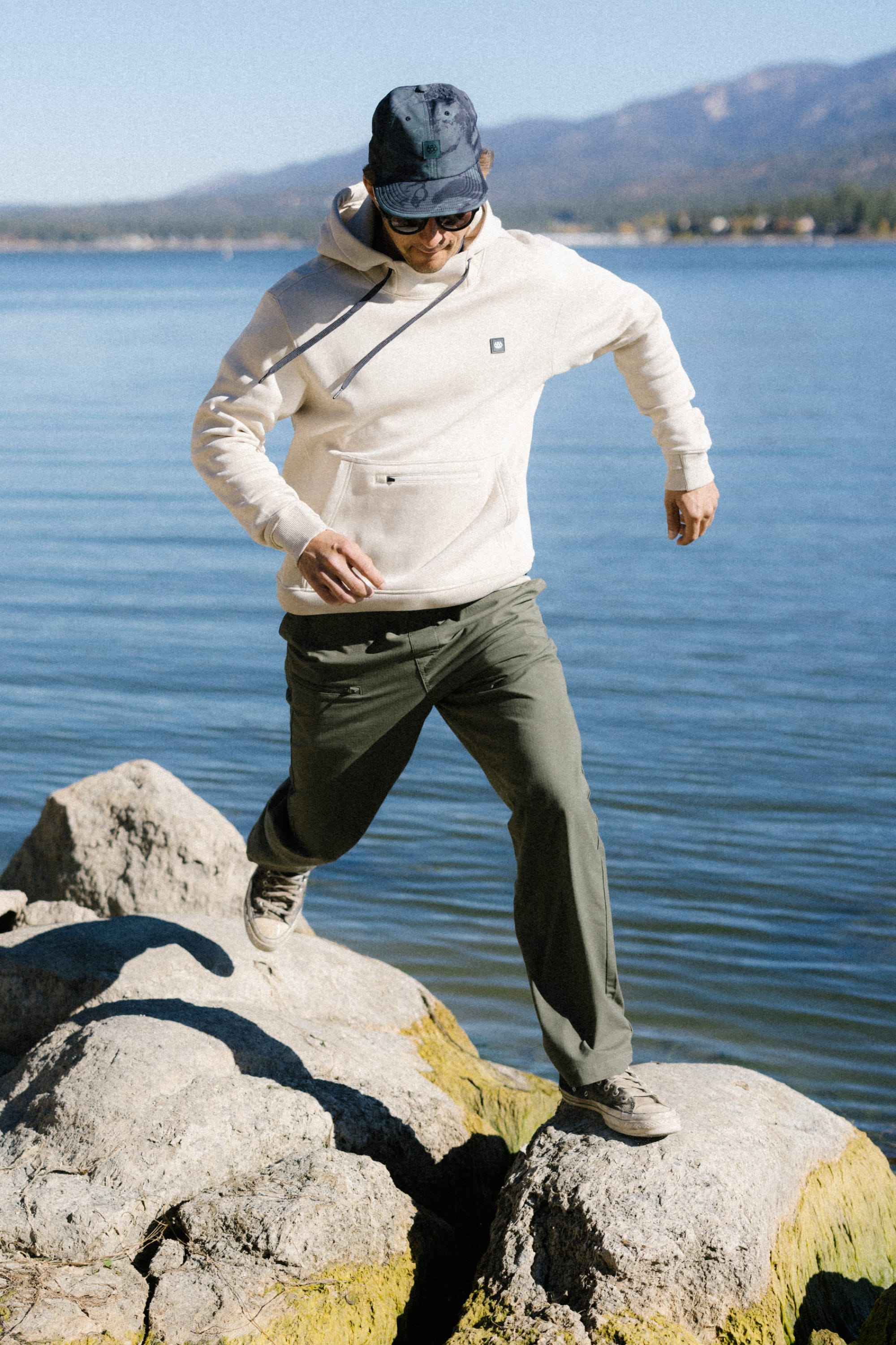 Wearing the 686 Men's All Season Hoody, a man—also sporting a cap and sunglasses—steps across rocks by a lake, with water and mountains in the background under clear skies.