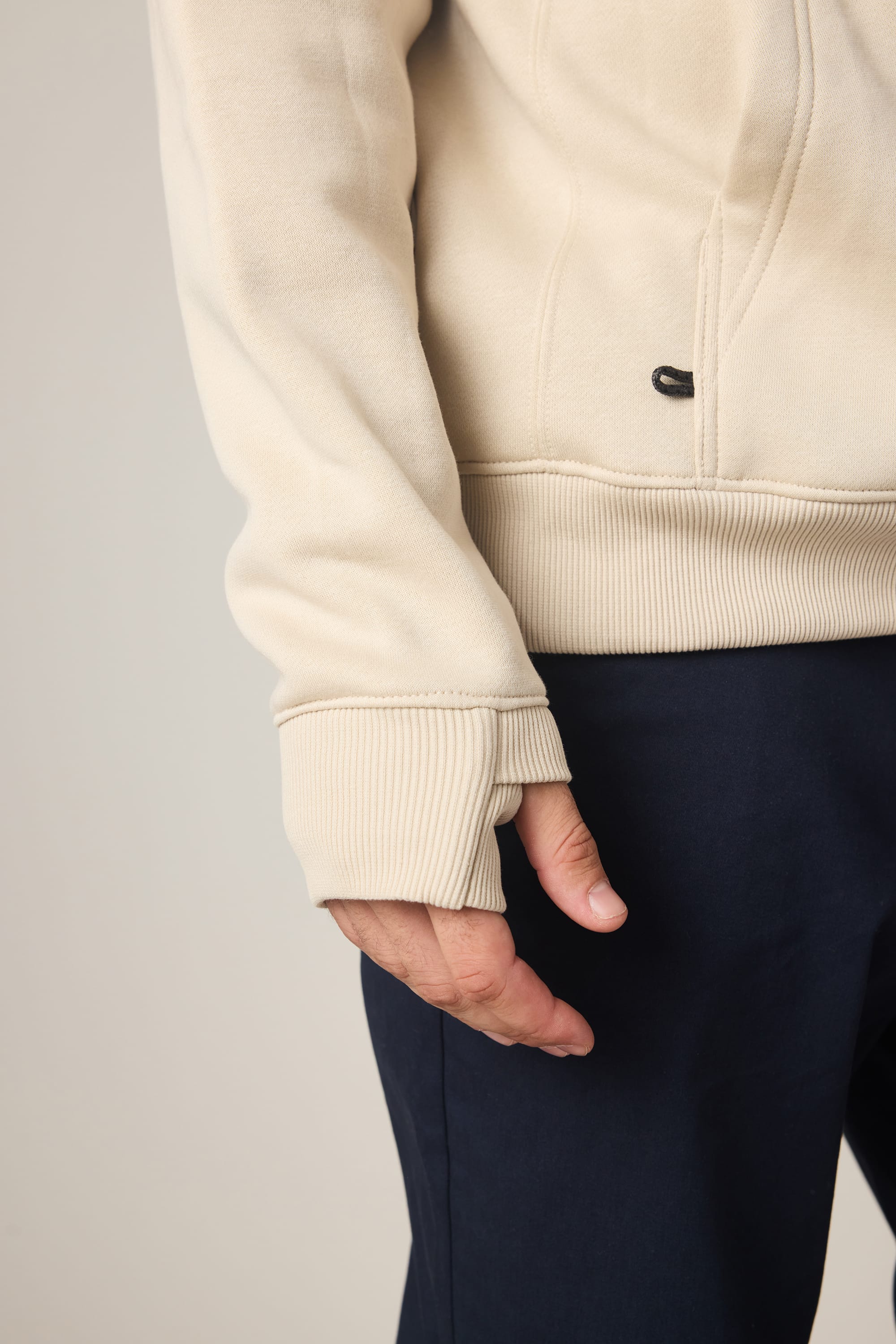 A person wearing the 686 Men's All Season Hoody in cream, made by 686, with ribbed cuffs and dark navy pants. Shown from the torso down with their left arm and hand visible.