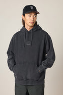 A man in the 686 Men's High Def Relaxed LS Hoody, featuring a small chest logo, black pants, and a black baseball cap stands with hands in the hoodie pocket, looking slightly to the side against a plain background.