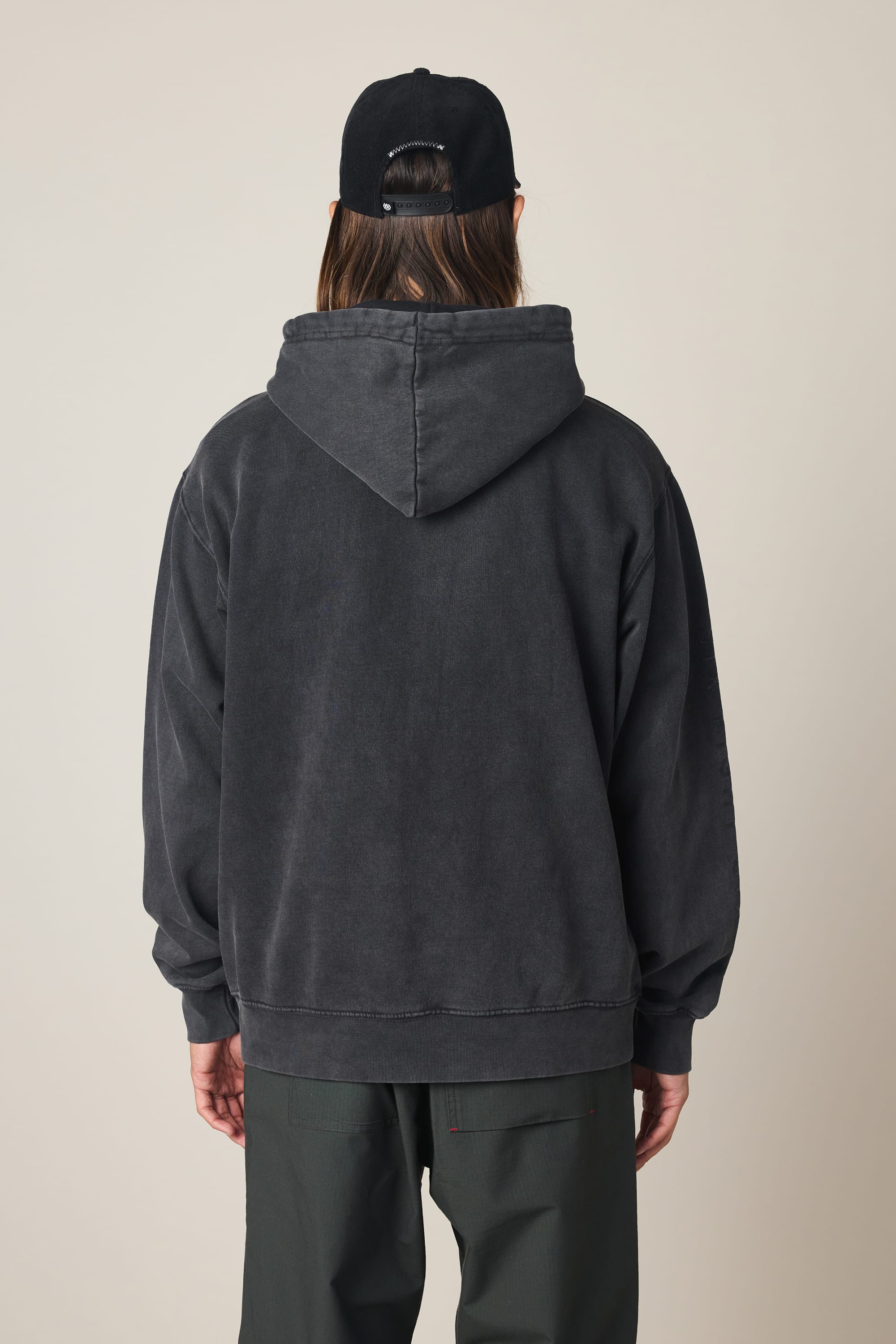 Wearing the 686 Men's High Def Relaxed LS Hoody in dark gray and a black baseball cap, a person with long hair stands with their back to the camera against a neutral background.