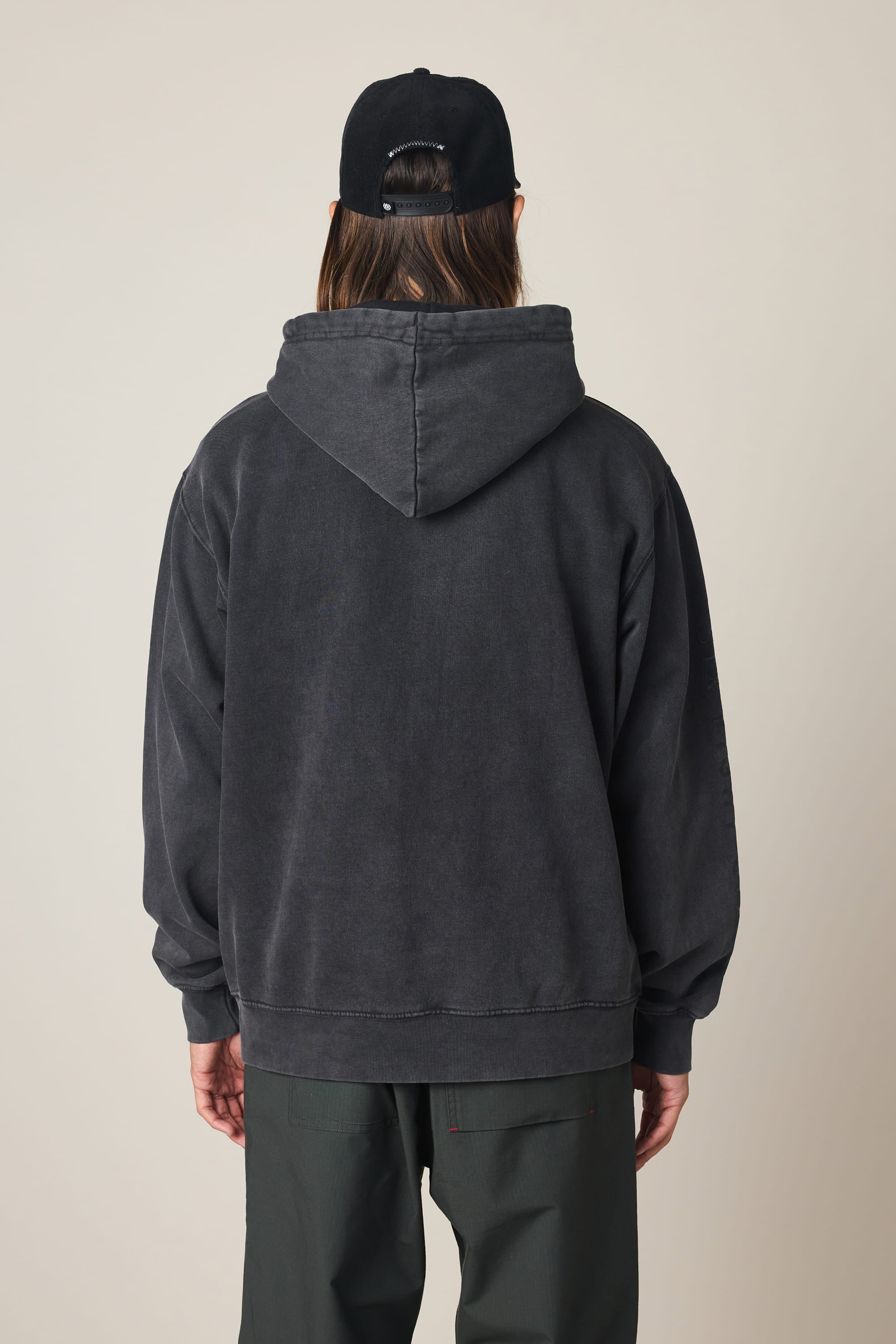 A person is standing with their back to the camera, wearing the 686 Men's High Def Relaxed LS Hoody in black, along with a black cap and dark pants. The hood of the 686 sweatshirt is down, covering most of their back and shoulders. The background is plain and light-colored.