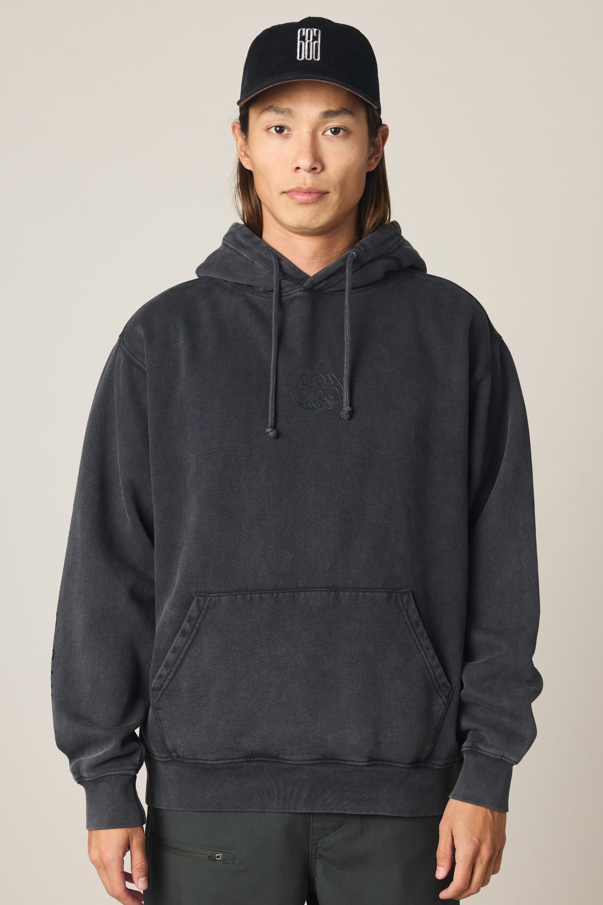 A person wearing the 686 Men's High Def Relaxed LS Hoody in black and a black baseball cap with white text stands facing forward against a plain, light-colored background.