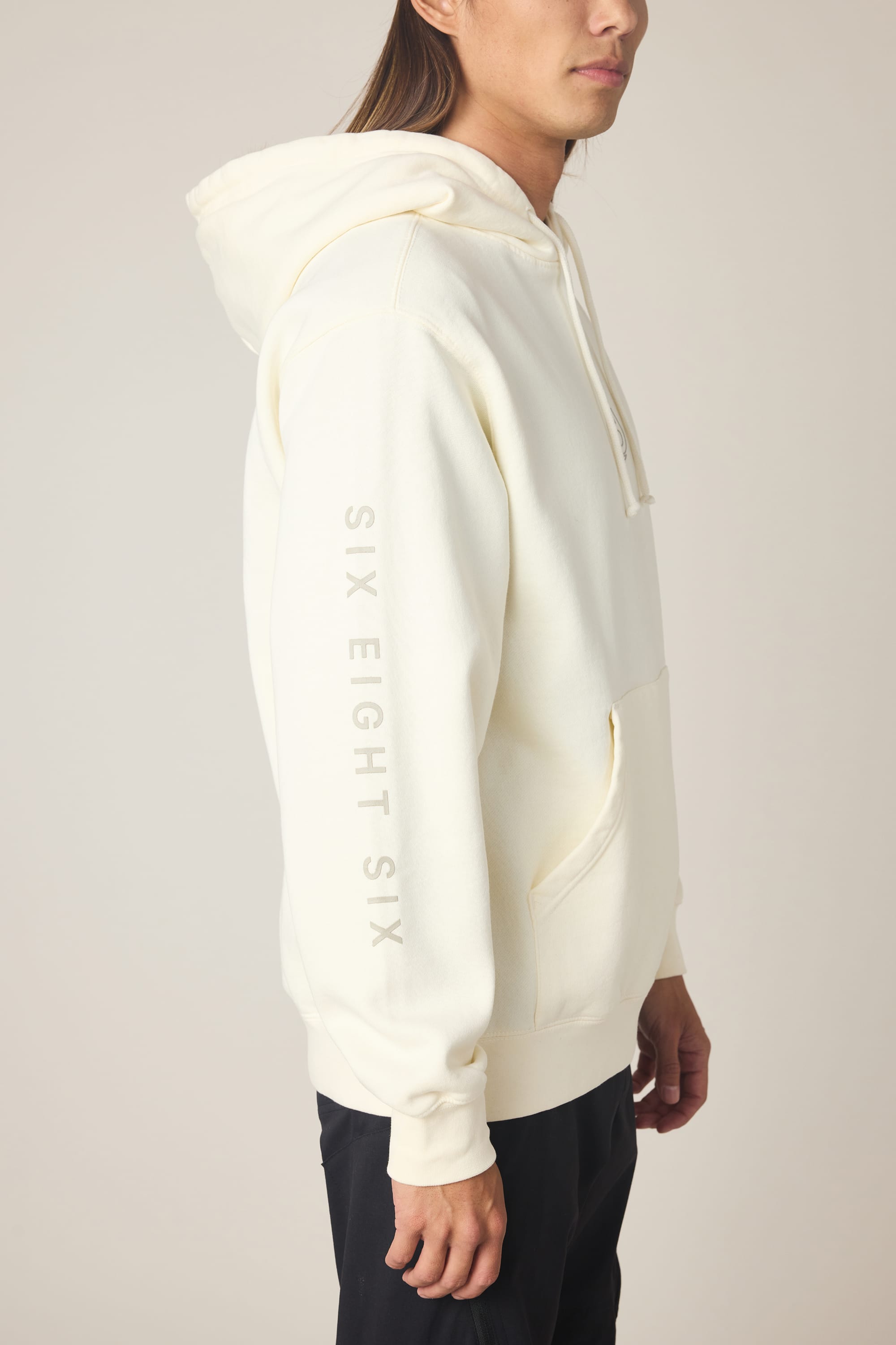 A person stands sideways with one hand by their side, wearing the 686 Men's High Def Relaxed LS Hoody in off-white, featuring light gray SIX EIGHT SIX lettering down the sleeve.