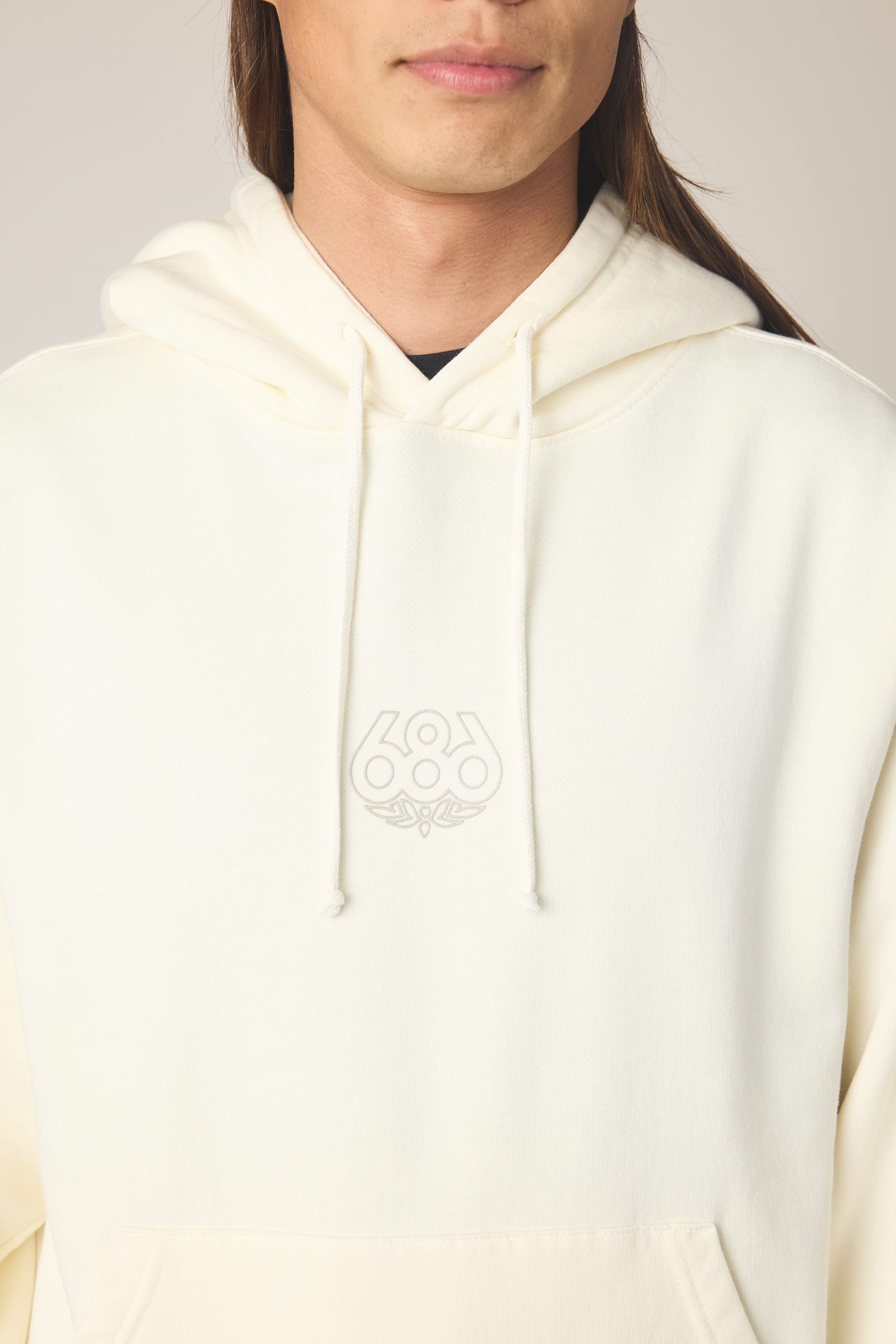 A person wears the 686 Men's High Def Relaxed LS Hoody, a cream-colored cotton sweatshirt with a 686 logo and chest design. Only the lower part of their face and long brown hair are visible.