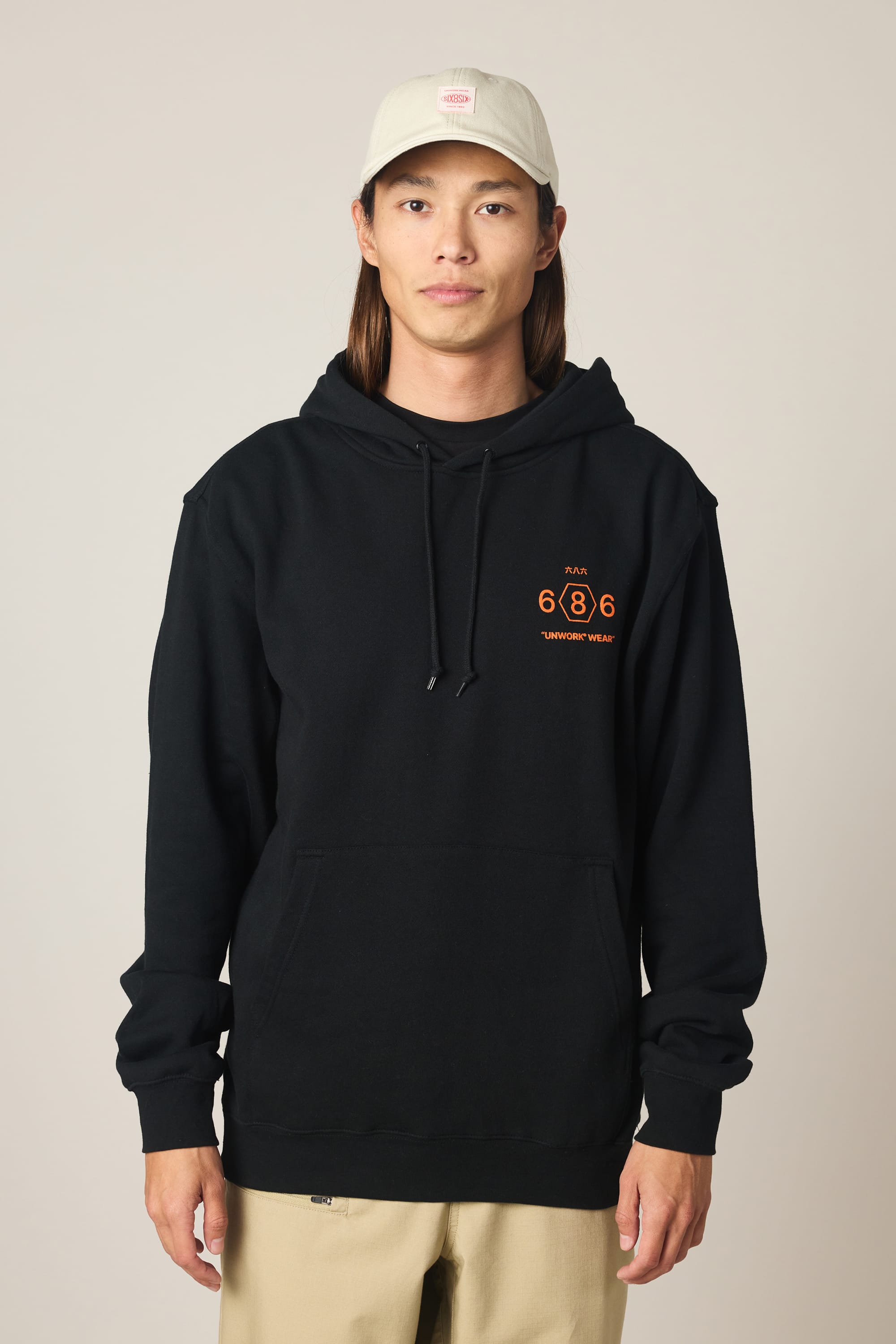 A person stands facing forward against a neutral background, wearing the 686 Men's Unwork Spec Athletic LS Hoody in black with orange graphics by 686, paired with a light-colored baseball cap and beige pants.