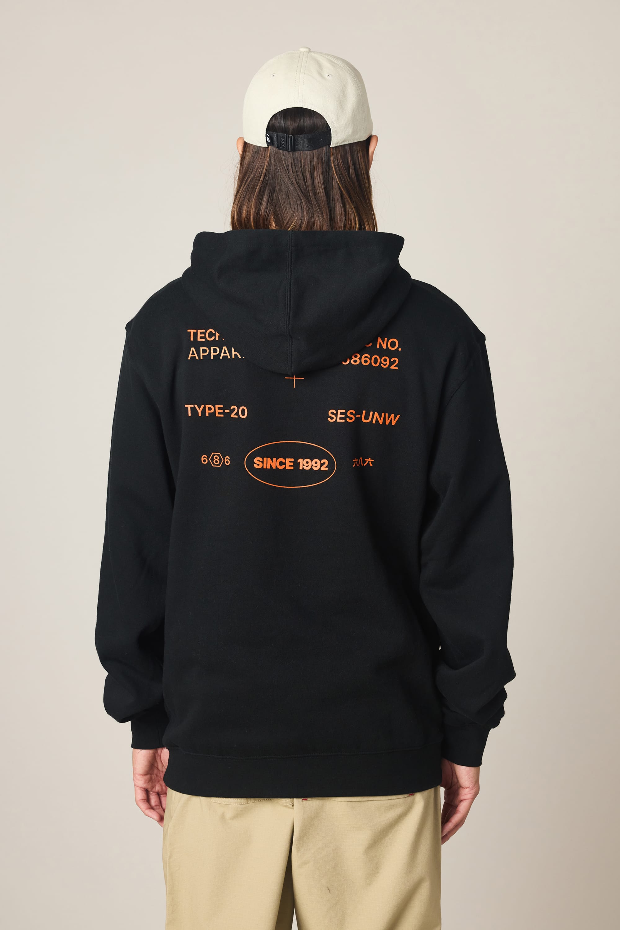 A person in the 686 Men's Unwork Spec Athletic LS Hoody—a black cotton sweatshirt with orange text and back graphics—faces away. They have long hair, a light baseball cap, and beige pants against a plain light background.