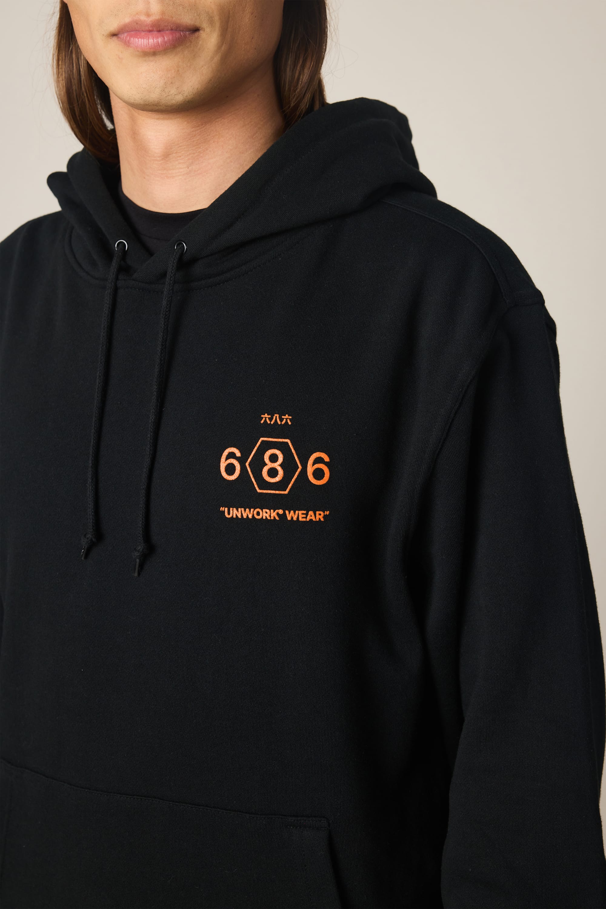 A person wears the 686 Men's Unwork Spec Athletic LS Hoody in black cotton, featuring orange "686" and "UNWORK WEAR" text and symbols. Only their lower face and upper torso are visible. Brand: 686.