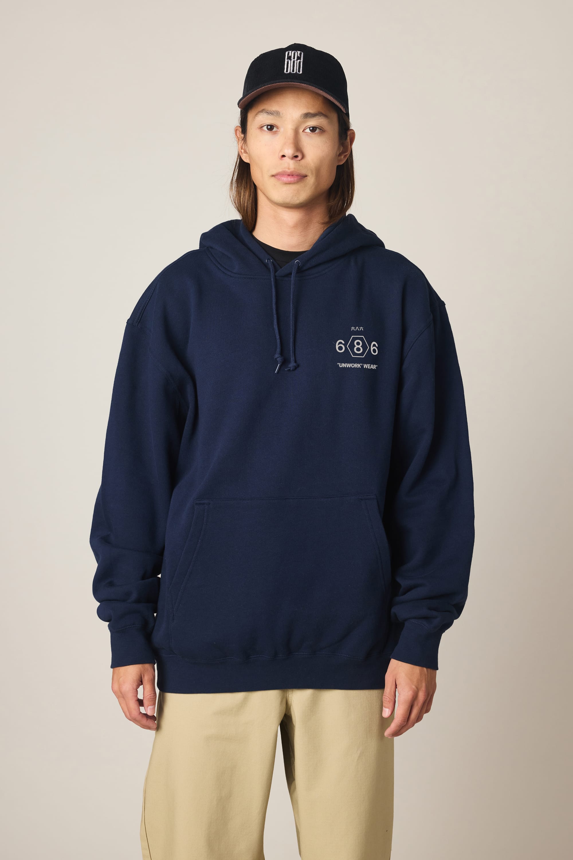 A person stands facing forward in a 686 Men's Unwork Spec Athletic LS Hoody (navy blue) with a small white logo and text on the left chest, beige pants, and a black baseball cap with a white design. The background is plain and light-colored.
