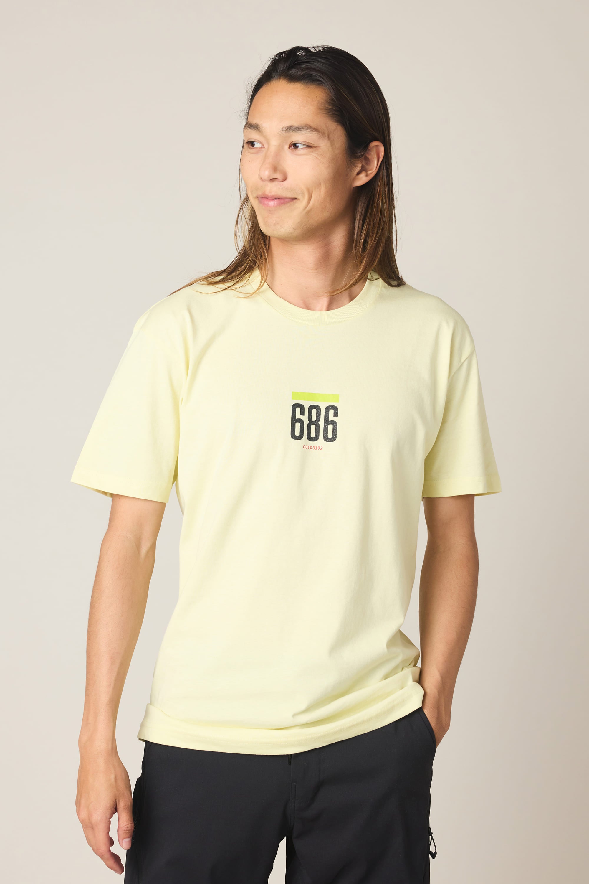 A person with long brown hair wears the 686 Lifts Authentic SS Tee by 686, a light yellow 100% cotton t-shirt with "686" on the front, paired with black pants, standing against a plain background and smiling to the side.