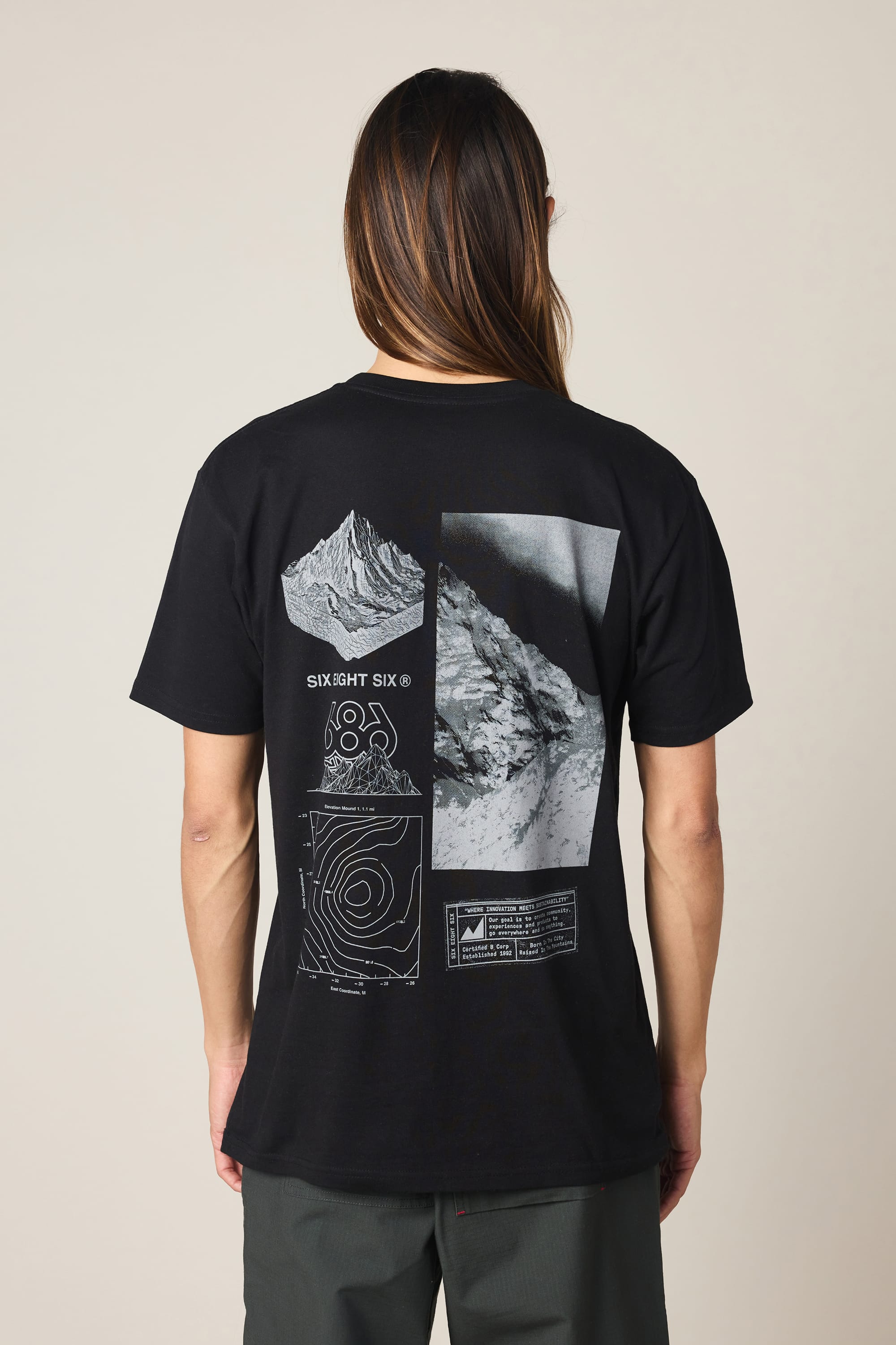 A person with long hair wears the 686 Cross-Cut Authentic S/S Tee by 686—a black men’s t-shirt featuring striking white graphic designs and mountain imagery on the back—standing against a plain light background. Made from 100% cotton, this tee delivers comfort and style for everyday wear.