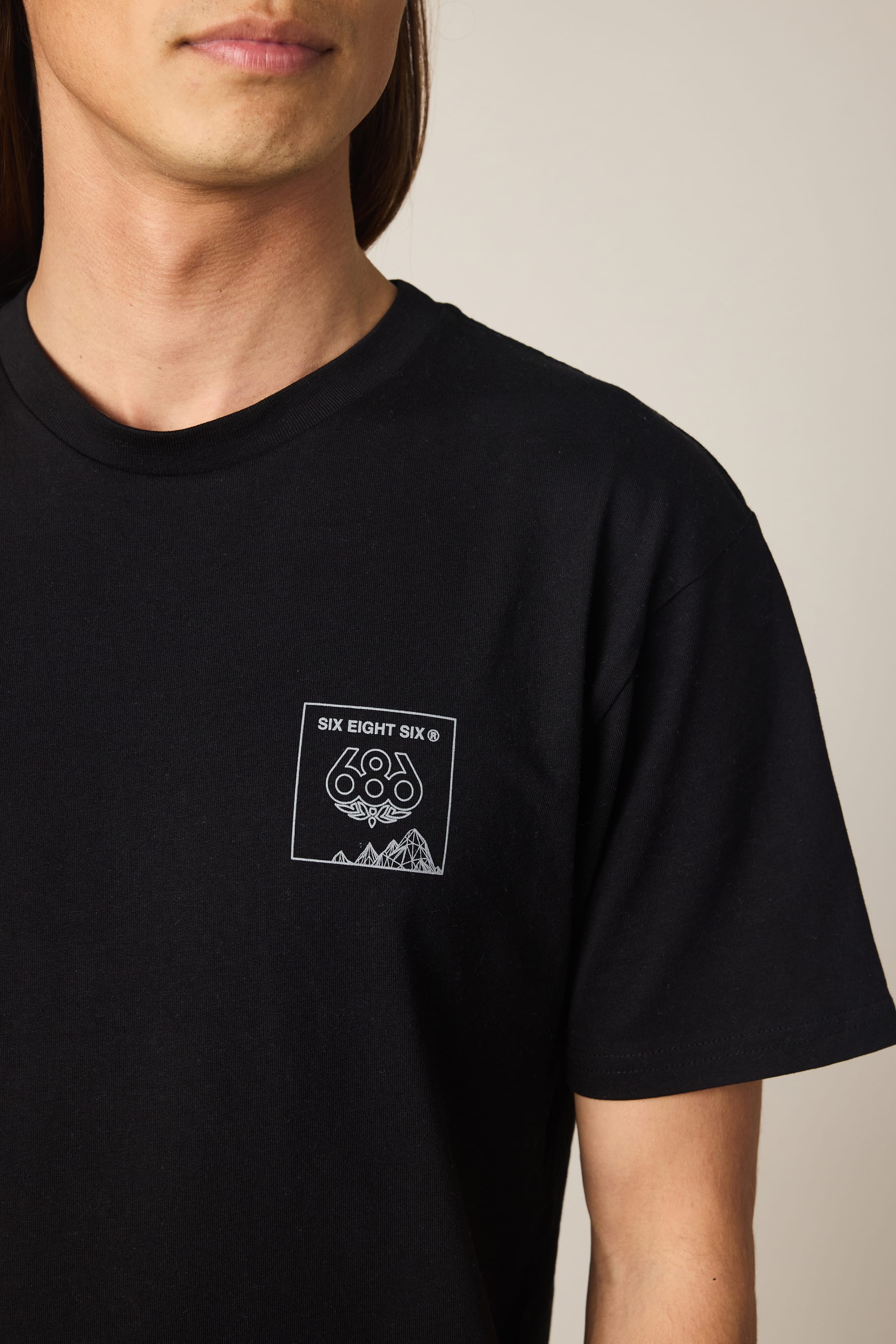 A person wearing the 686 Cross-Cut Authentic S/S Tee by 686—a men's T-shirt in black, made from 100% cotton, featuring a small white graphic and the text "SIX EIGHT SIX" on the chest. The image partially shows the person's head and highlights this classic tee design.