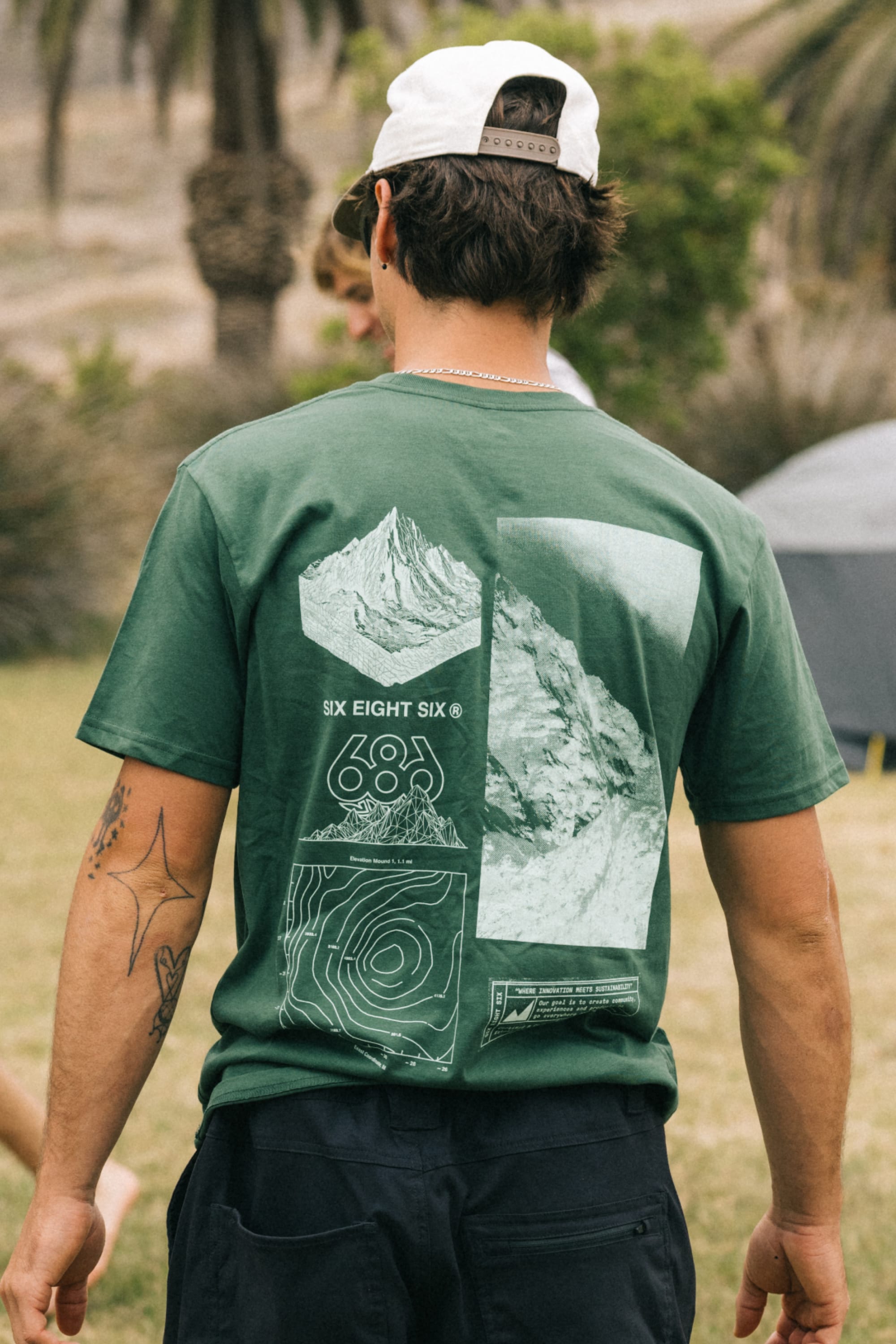 Wearing the 686 Cross-Cut Authentic S/S Tee by 686, a person with their back to the camera stands outdoors in dark shorts and a white backwards baseball cap near trees and a tent. The green tee is made from 100% cotton and features mountain graphics.