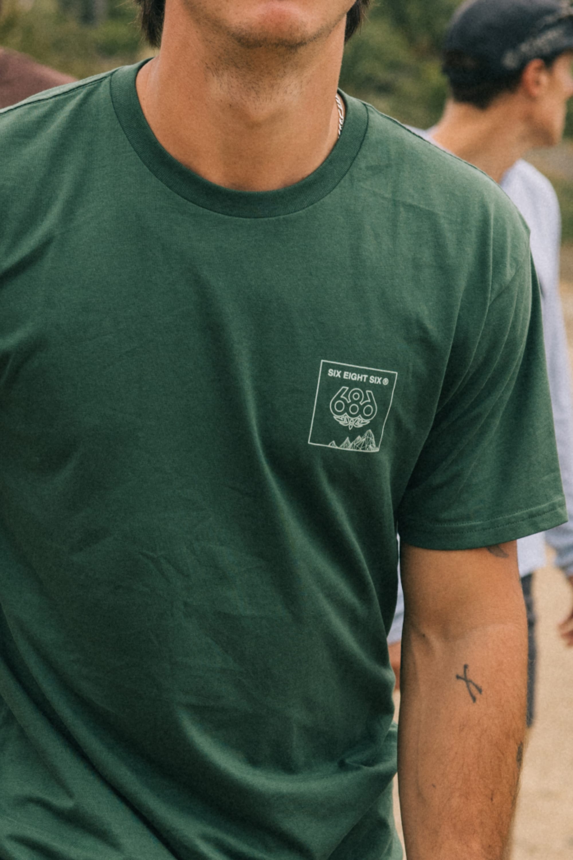 A person stands outdoors wearing the 686 Cross-Cut Authentic S/S Tee by 686, a green mens cotton t-shirt with a small geometric chest design. In the background, another person in a cap and light shirt appears slightly blurred.
