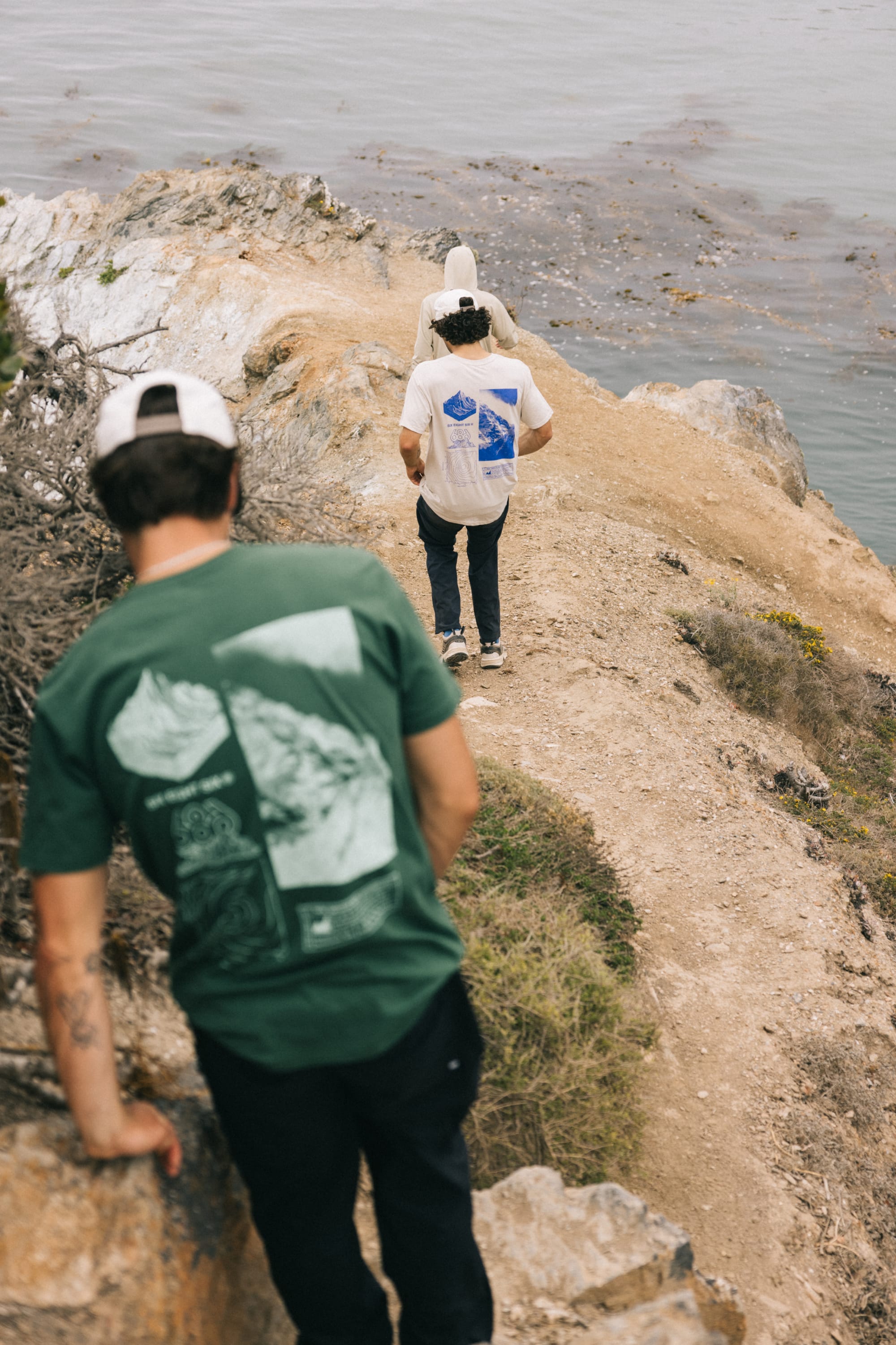 Two people in 686 Cross-Cut Authentic S/S Tees walk along a rocky, narrow ocean path. One leads while the other follows, both framed by scenic coastal water and rocks in the background.