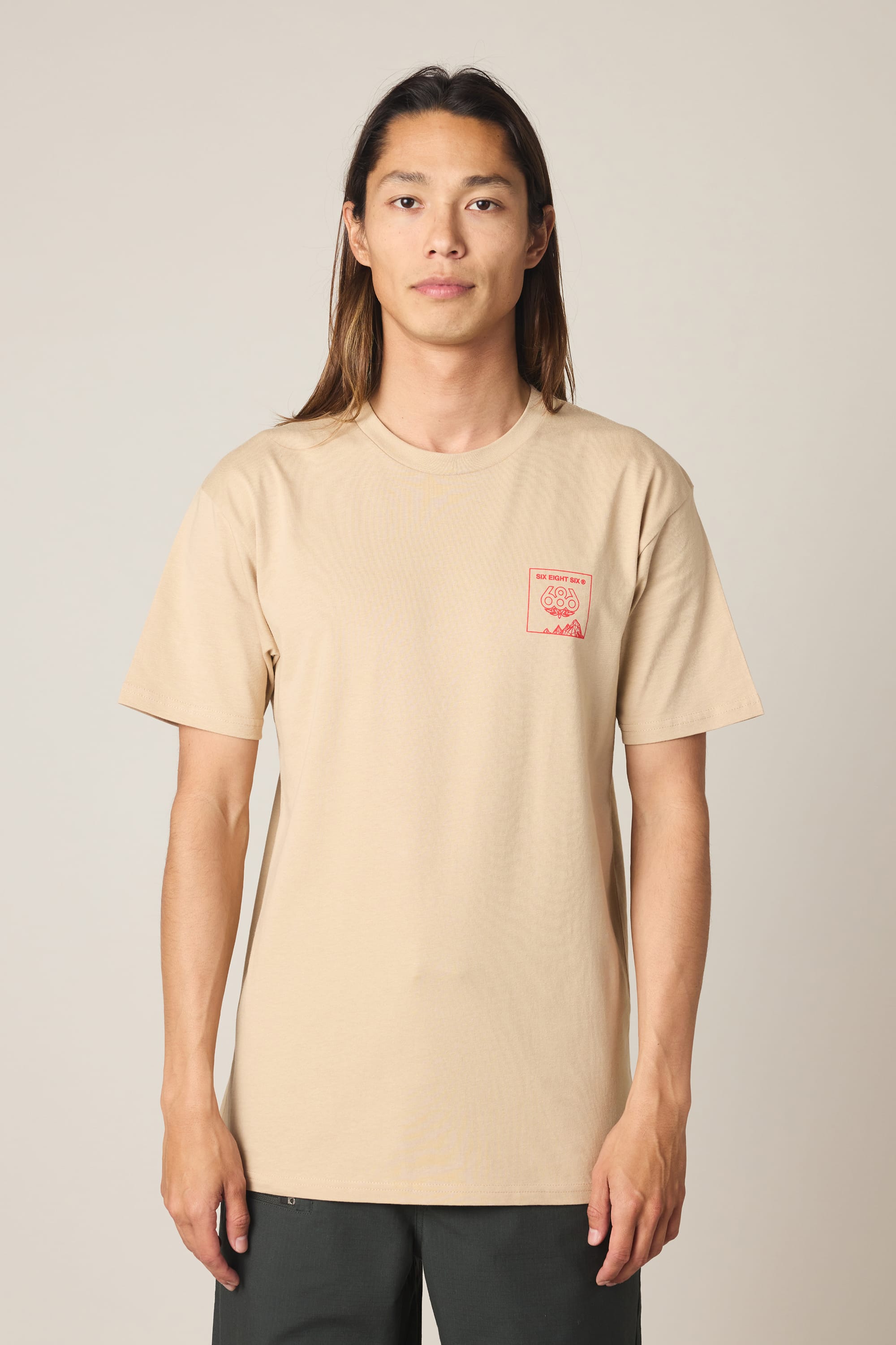A person with long hair wears the 686 Cross-Cut Authentic S/S Tee by 686—a beige men’s t-shirt crafted from 100% cotton, featuring a small red logo and text on the left chest—while standing against a plain light background.