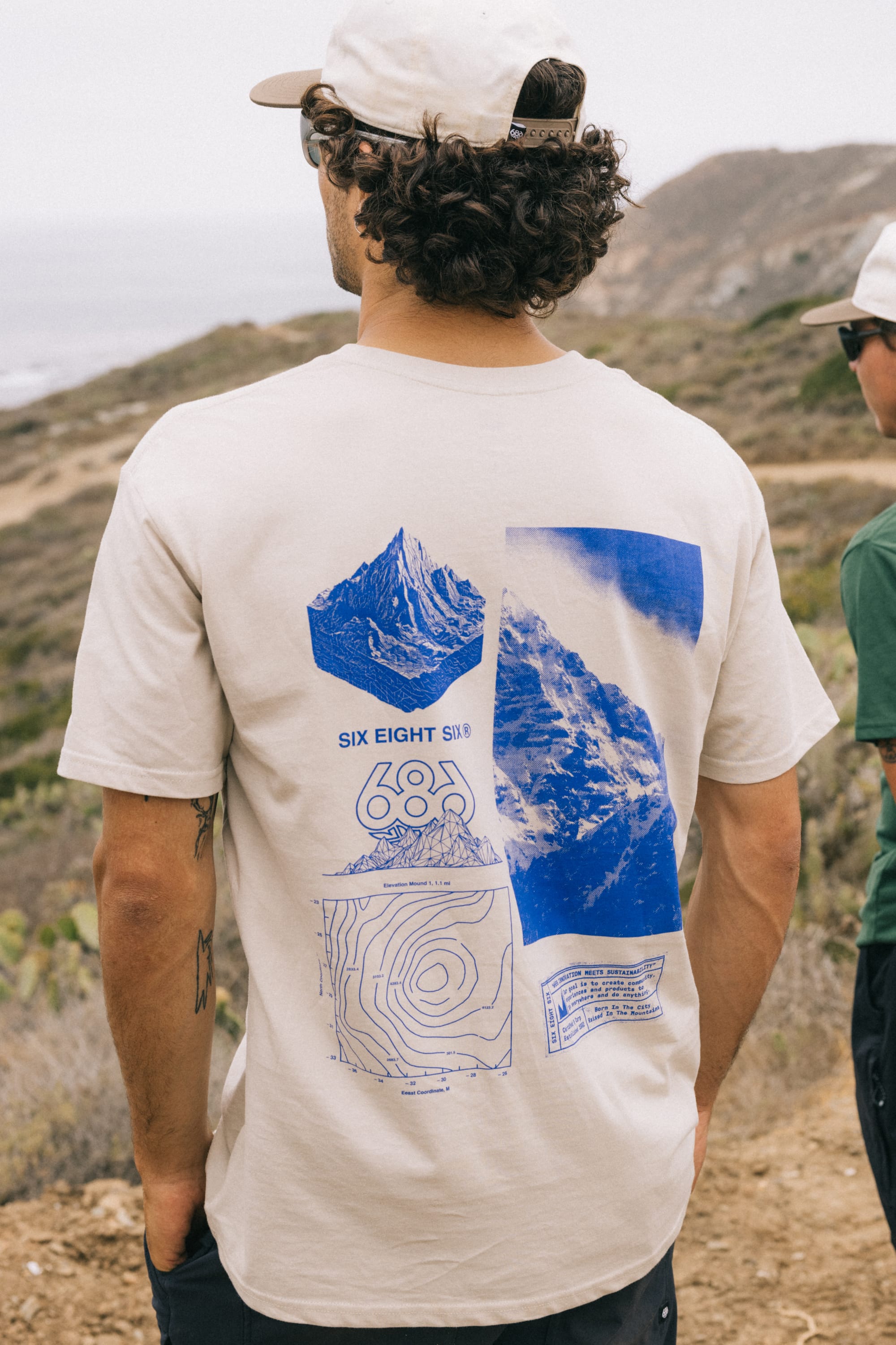 A man wears the 686 Cross-Cut Authentic S/S Tee in beige, featuring blue mountain graphics and SIX EIGHT SIX text, while standing outdoors by the coast. Another person in a green shirt and white cap is partially visible.