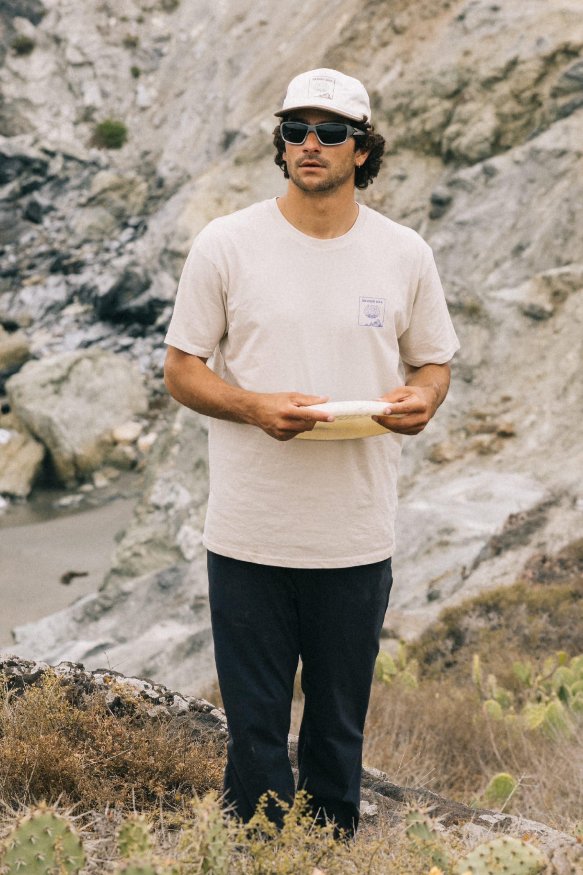 A man in sunglasses, a white cap, and a beige 686 Cross-Cut Authentic S/S Tee by 686 stands on rocky terrain holding a frisbee, surrounded by dried grass and cliffs.