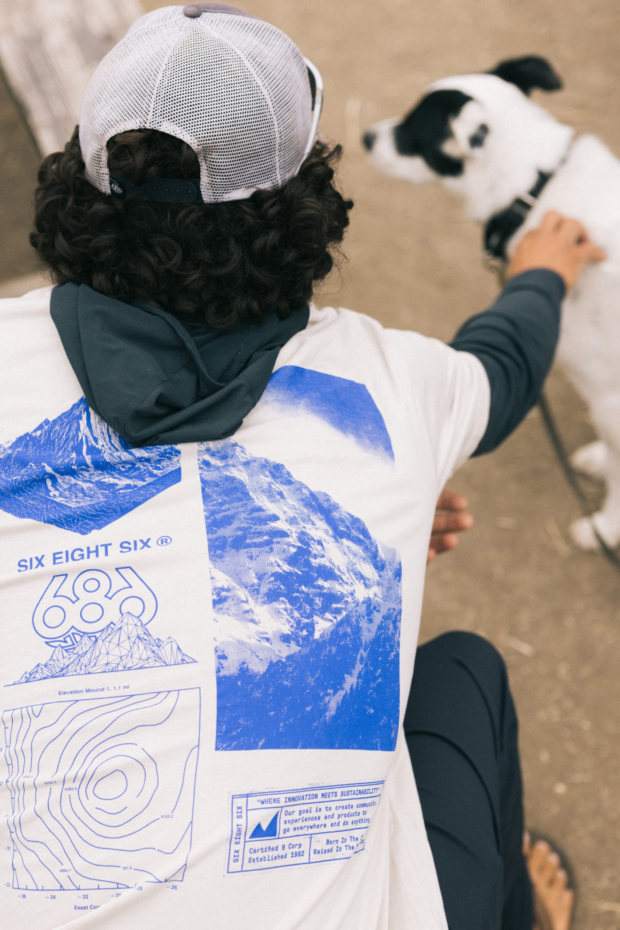 A person with curly hair in a baseball cap wears the 686 Cross-Cut Authentic S/S Tee by 686, which features blue mountain and map designs, while petting a black and white dog outdoors.
