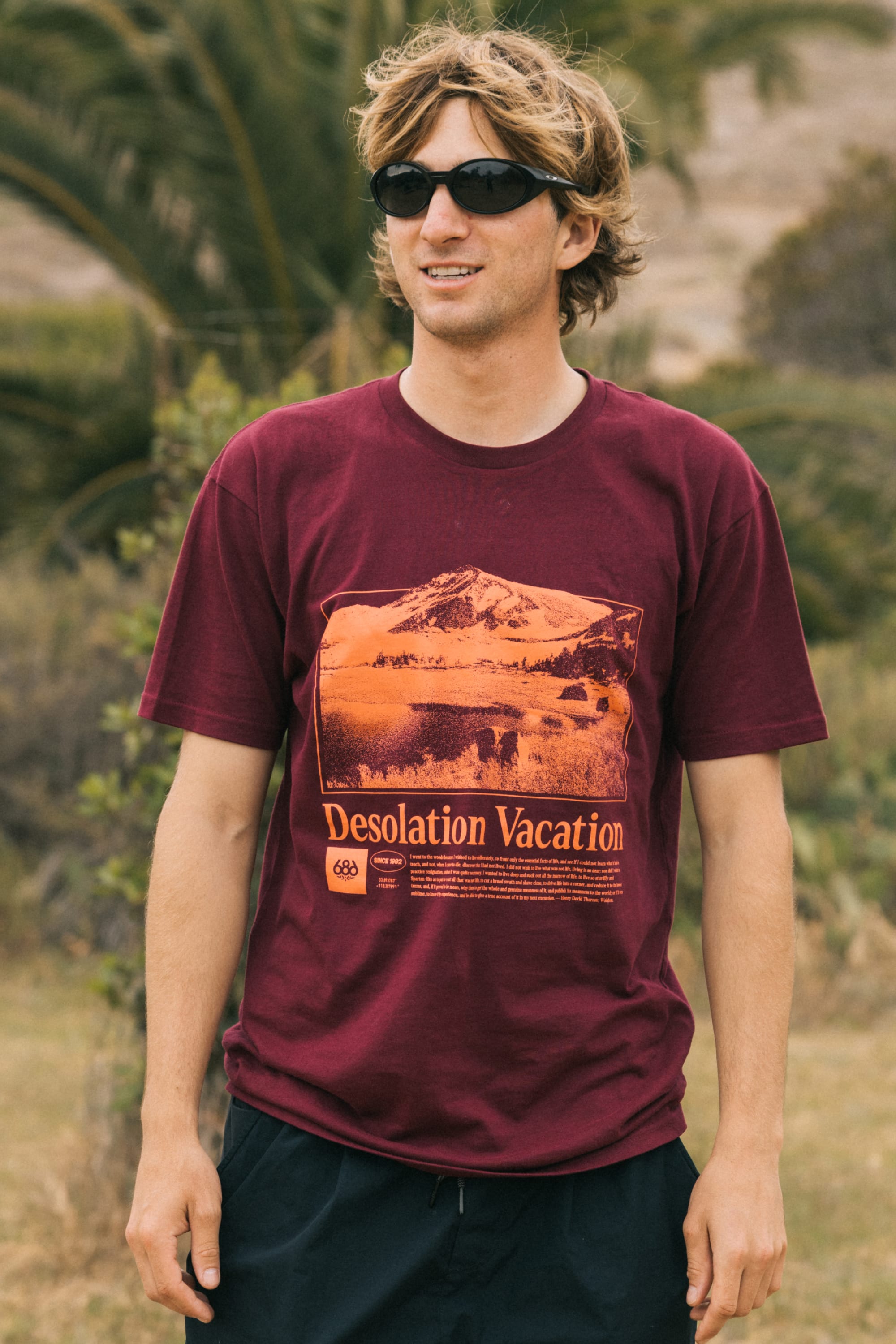 A man with light brown hair and black sunglasses wears the 686 Desolation Vacation Authentic S/S Tee and black shorts, standing outdoors with greenery and a palm tree in the background.
